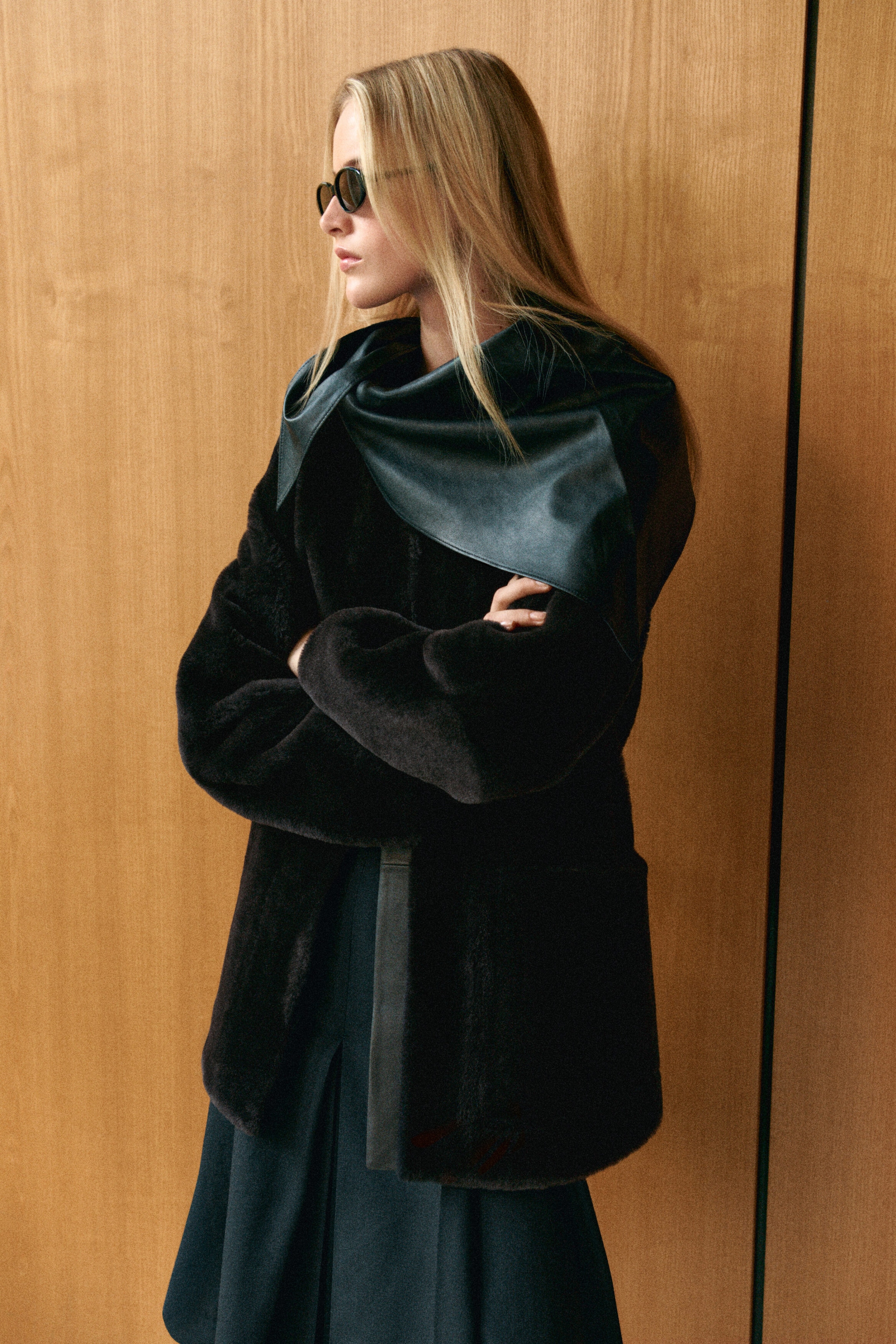 This collarless shearling jacket in dark brown is shown from a three-quarter front angle, featuring a plush, textured exterior and a smooth leather trim along the front placket. It is styled with a draped black leather neckpiece layered over the shoulders, accentuating its collarless neckline. This collarless shearling jacket in dark brown is shown from a three-quarter front angle, featuring a plush, textured exterior and a smooth leather trim along the front placket. It is styled with a draped black leather neckpiece layered over the shoulders, accentuating its collarless neckline.