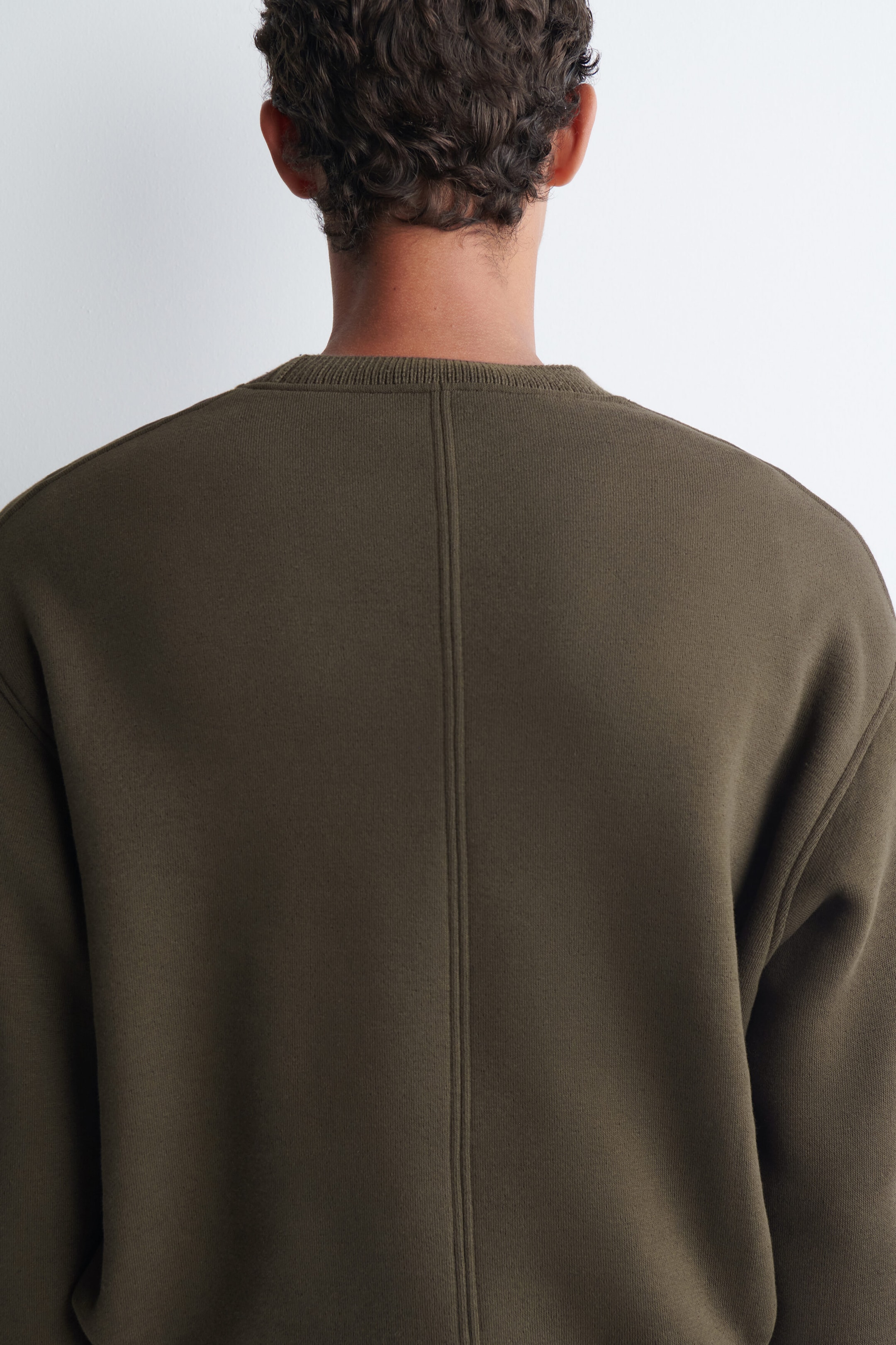 Un chandail en molleton de coton de couleur vert olive foncé est porté par le mannequin, présenté de dos. Le vêtement présente un col côtelé et une couture verticale distinctive qui descend au centre du dos. Un chandail en molleton de coton de couleur vert olive foncé est porté par le mannequin, présenté de dos. Le vêtement présente un col côtelé et une couture verticale distinctive qui descend au centre du dos.