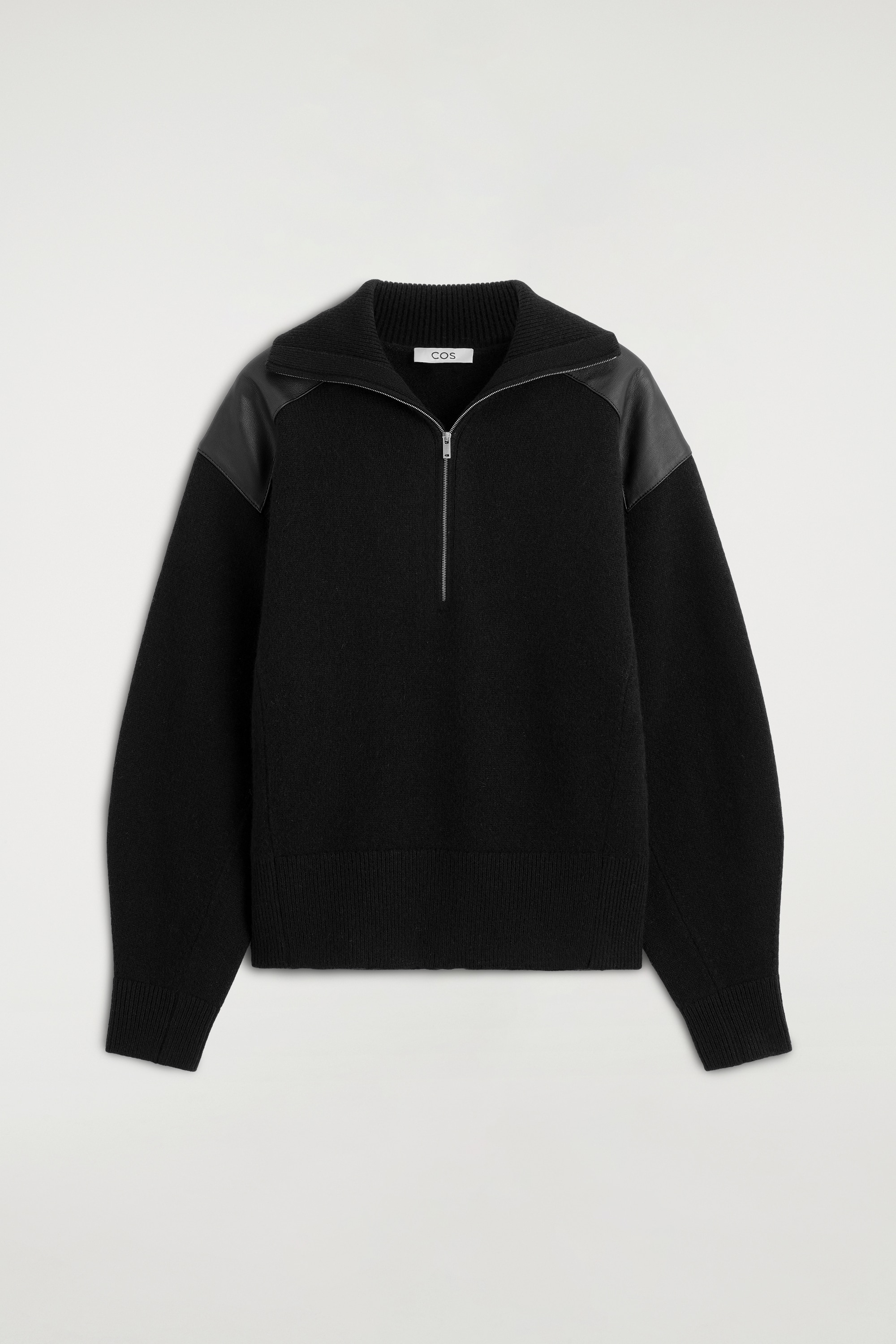 A modelo estÃ¡ a usar uma camisola de merino com fecho de correr e aplicaÃ§Ã£o em pele preta, com gola alta e fecho de meio fecho em tom prateado, e painÃ©is em pele tonal nos ombros. A camisola estÃ¡ metida por dentro de umas calÃ§as pretas de cintura subida, complementadas por um cinto fino branco.