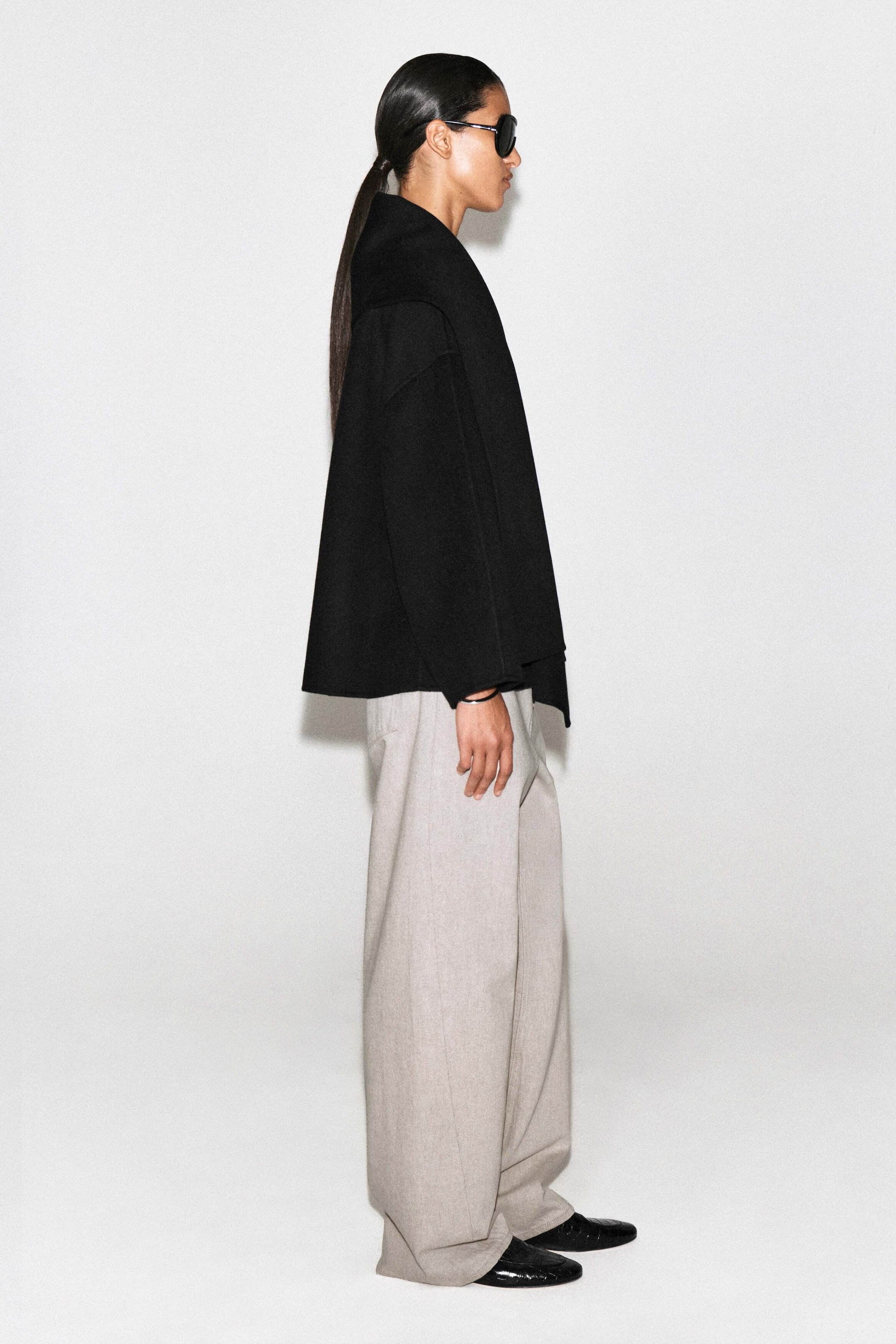 The double-faced wool-blend scarf jacket in black features a loose, boxy silhouette in a right-facing pose, highlighting its integrated scarf collar draped over the shoulder. Falling to mid-hip length, the jacket is styled against light-colored wide-leg trousers to emphasize its structured drape.