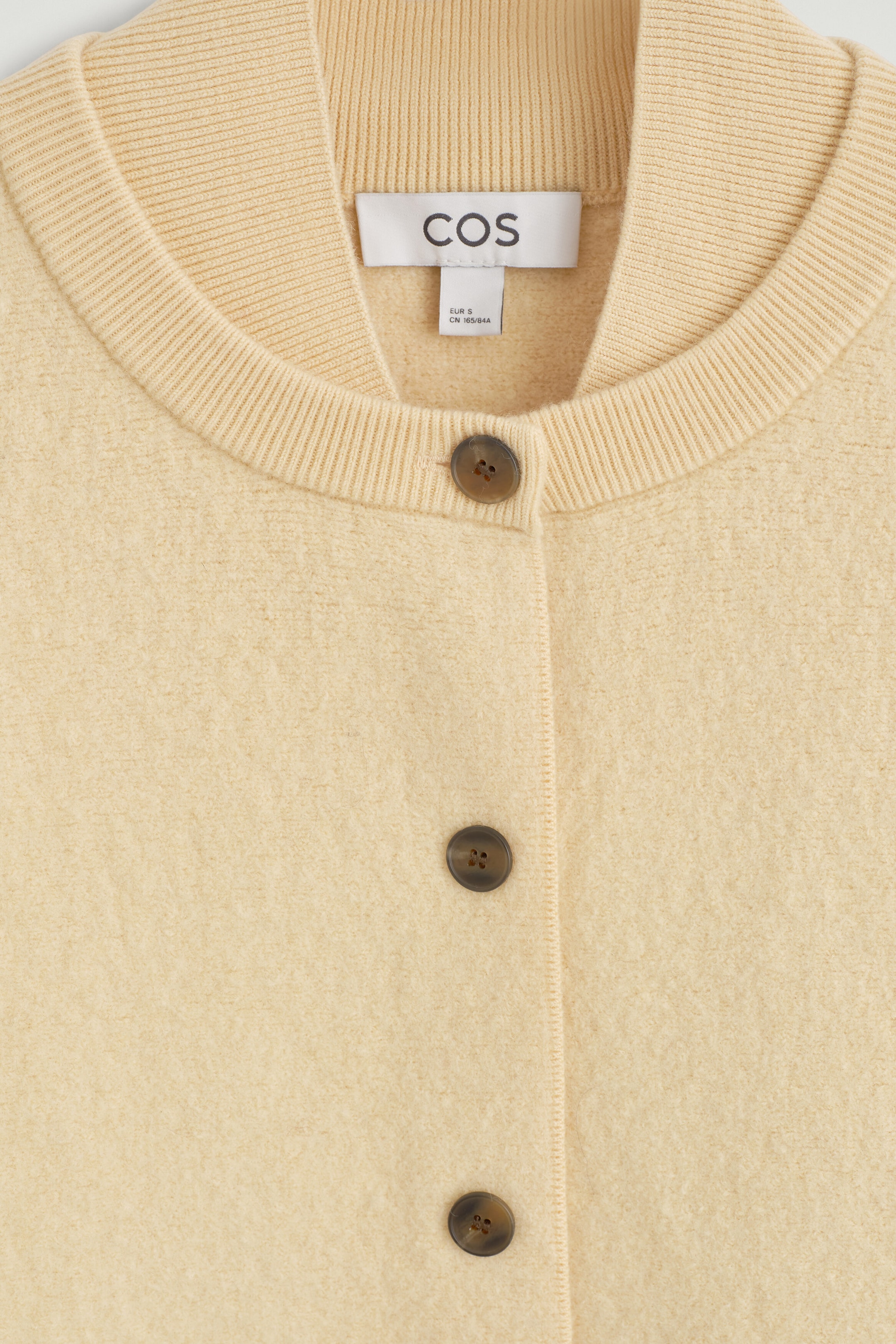 Une vue rapprochée de face d’un cardigan en laine bouillie jaune pâle mettant en valeur sa finition texturée, son col côtelé à double épaisseur et ses boutons brun foncé sur la patte de boutonnage à l’avant. Une vue rapprochée de face d’un cardigan en laine bouillie jaune pâle mettant en valeur sa finition texturée, son col côtelé à double épaisseur et ses boutons brun foncé sur la patte de boutonnage à l’avant.