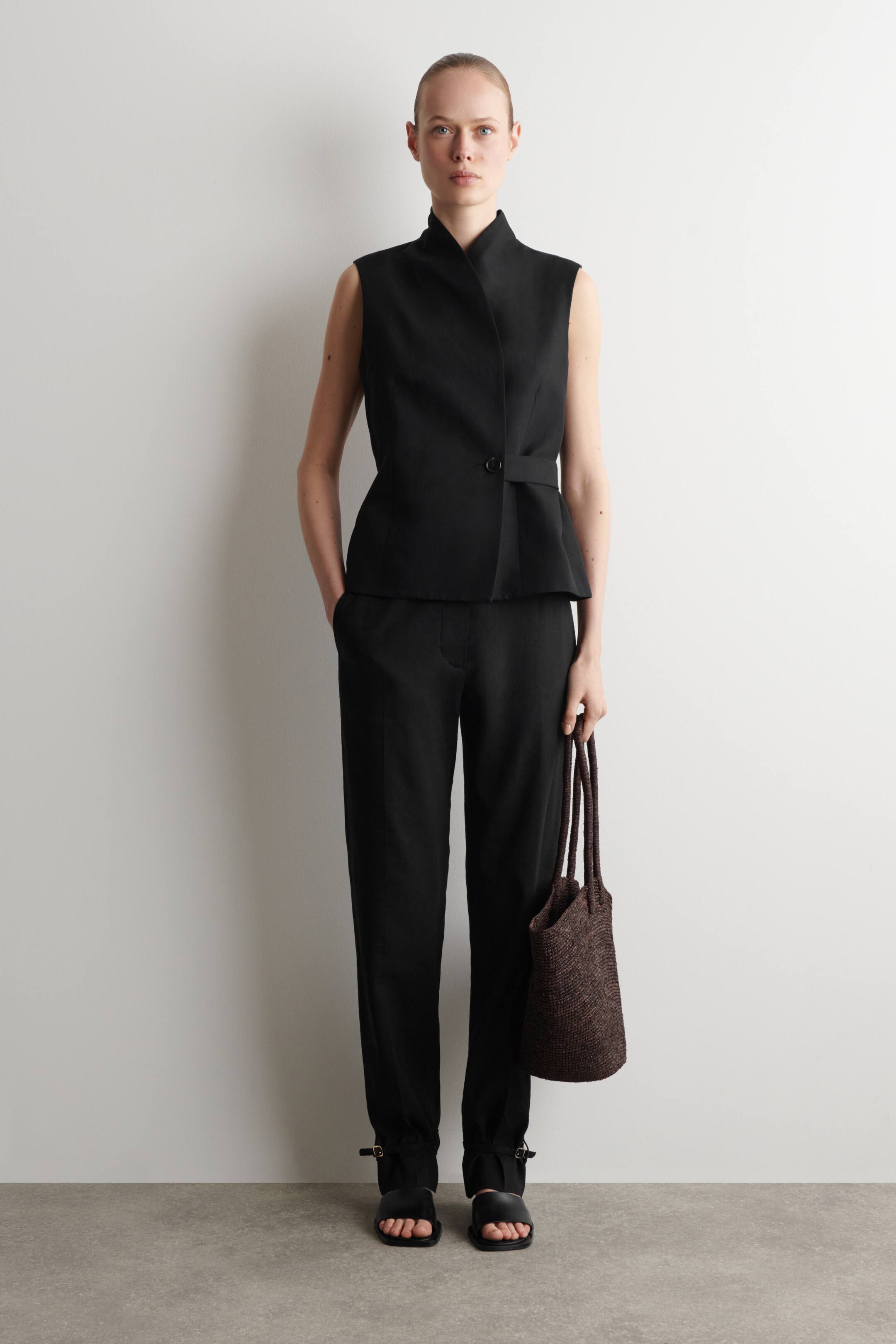 Ver imagem maior: Um top preto sem mangas, virado para a frente, apresenta uma gola alta e sobreposta, um fecho com botão único na cintura com uma aba lateral e uma ligeira bainha peplum. Calças pretas acompanham o top, exibindo pernas retas e alças com fivelas nos tornozelos.