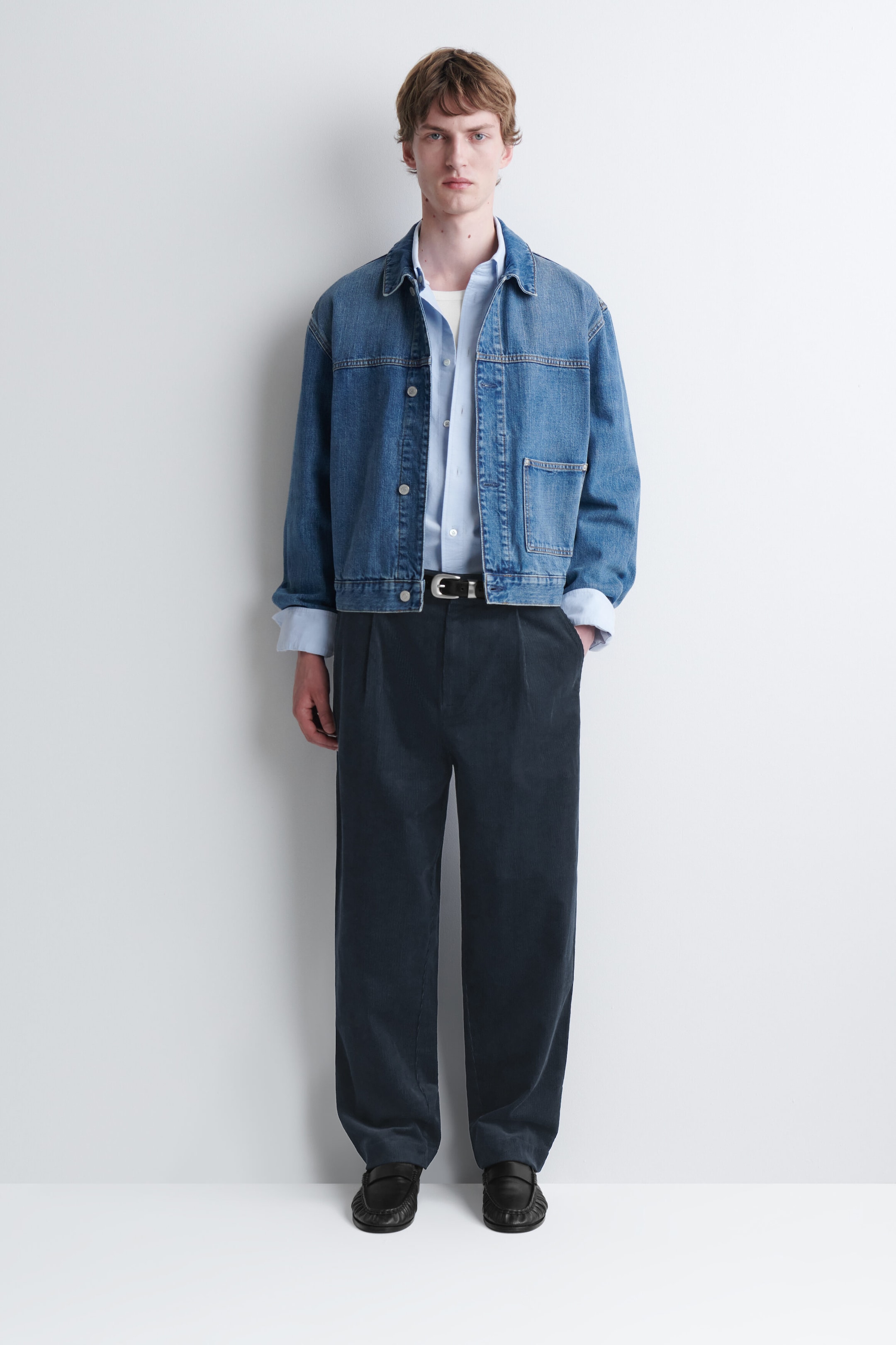 Corduroy relaxed-fit tapered pants in dark navy blue are being worn by the model, who is standing with one hand in a pocket. The pleated pants are styled with a black leather belt, a light blue button-up shirt, a blue denim jacket, and black leather loafers. Corduroy relaxed-fit tapered pants in dark navy blue are being worn by the model, who is standing with one hand in a pocket. The pleated pants are styled with a black leather belt, a light blue button-up shirt, a blue denim jacket, and black leather loafers.