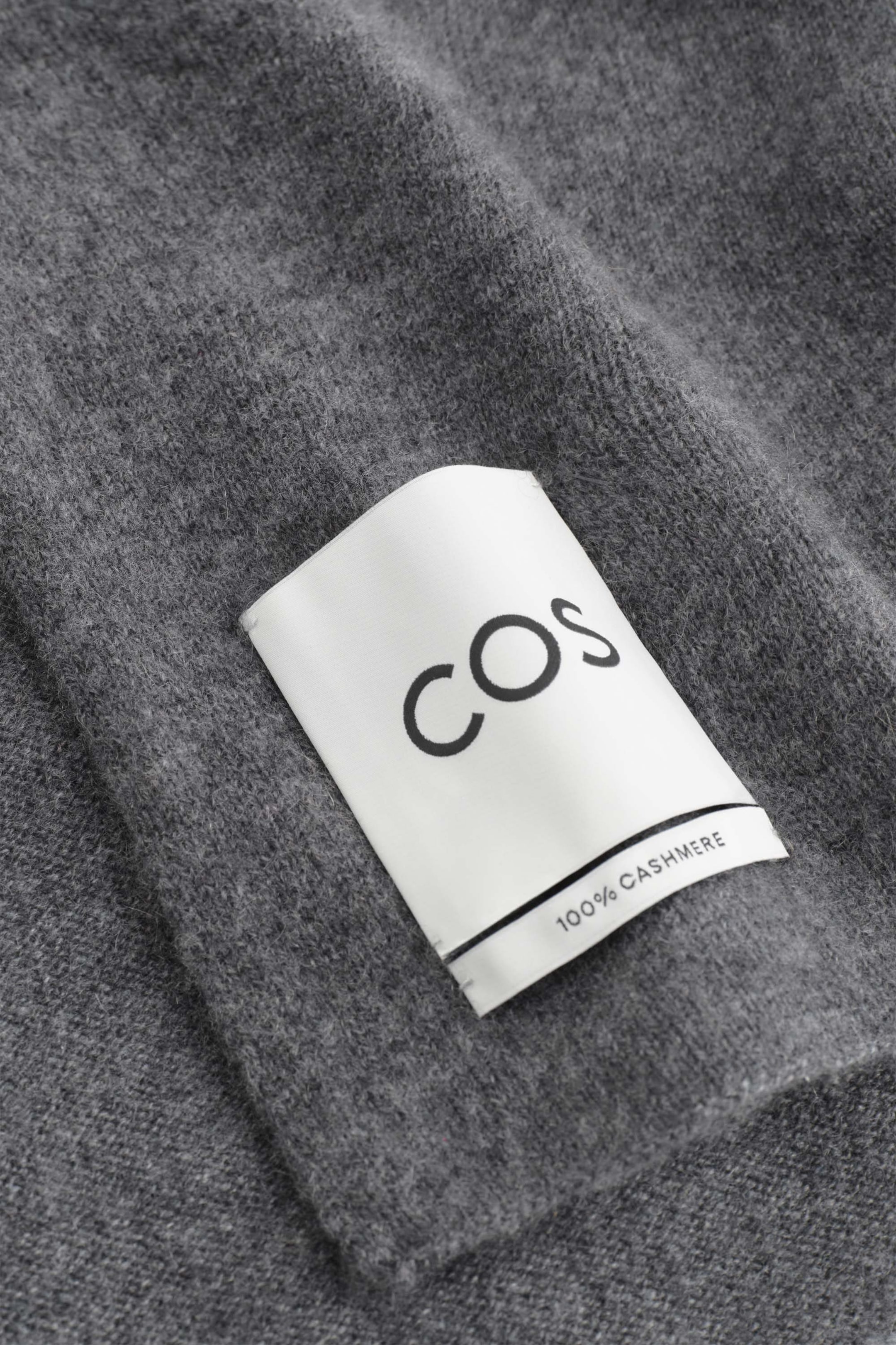 A grey cashmere scarf is shown in close-up, highlighting its soft, textured knit. A white fabric label is stitched onto the fabric, displaying the brand name "COS" and "100% CASHMERE" in black lettering. A grey cashmere scarf is shown in close-up, highlighting its soft, textured knit. A white fabric label is stitched onto the fabric, displaying the brand name "COS" and "100% CASHMERE" in black lettering.