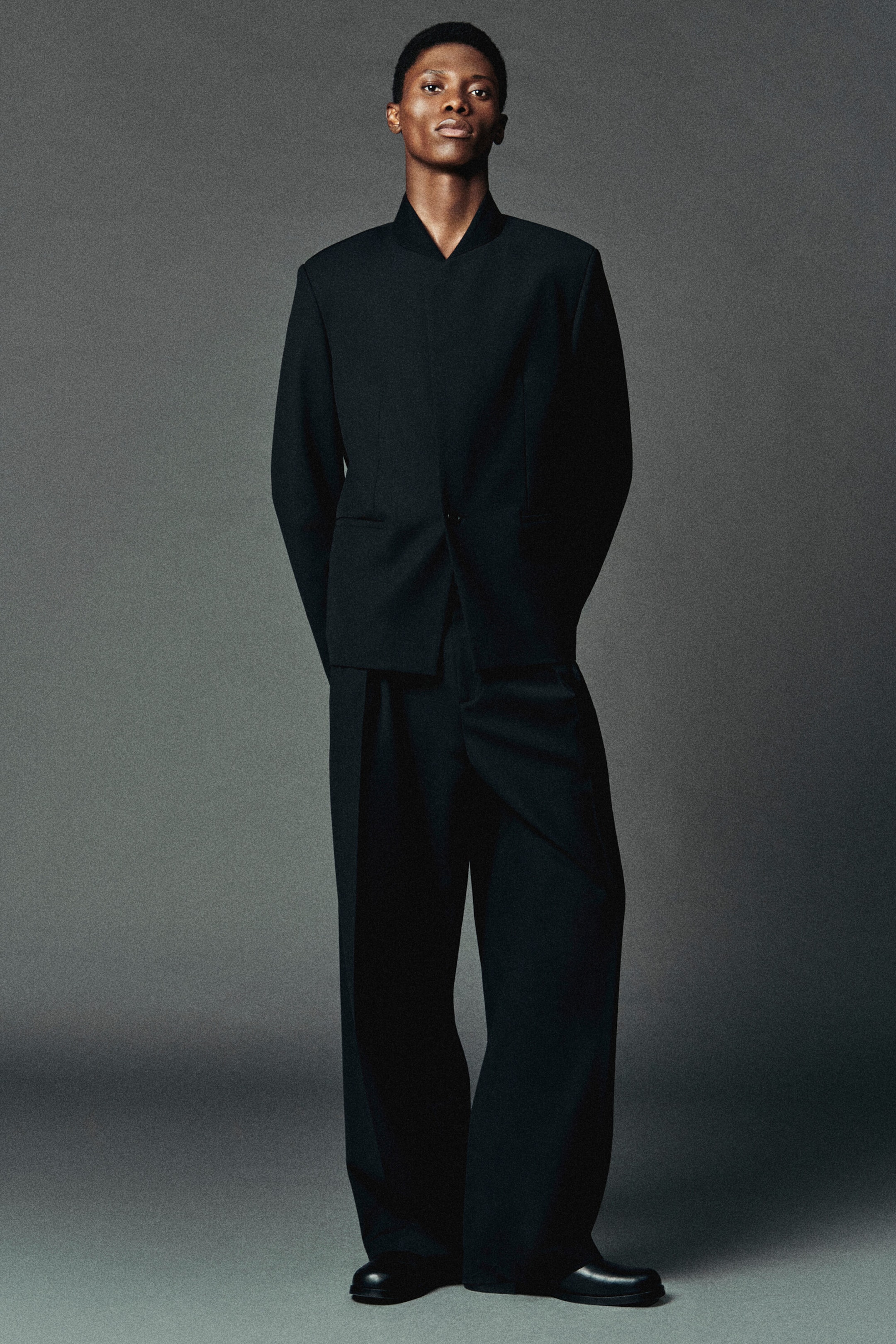 This black wool-blend blazer, featuring a structured stand collar and asymmetric fastening, is shown from the front paired with matching black relaxed-fit wool trousers that have pleated barrel legs. This black wool-blend blazer, featuring a structured stand collar and asymmetric fastening, is shown from the front paired with matching black relaxed-fit wool trousers that have pleated barrel legs.
