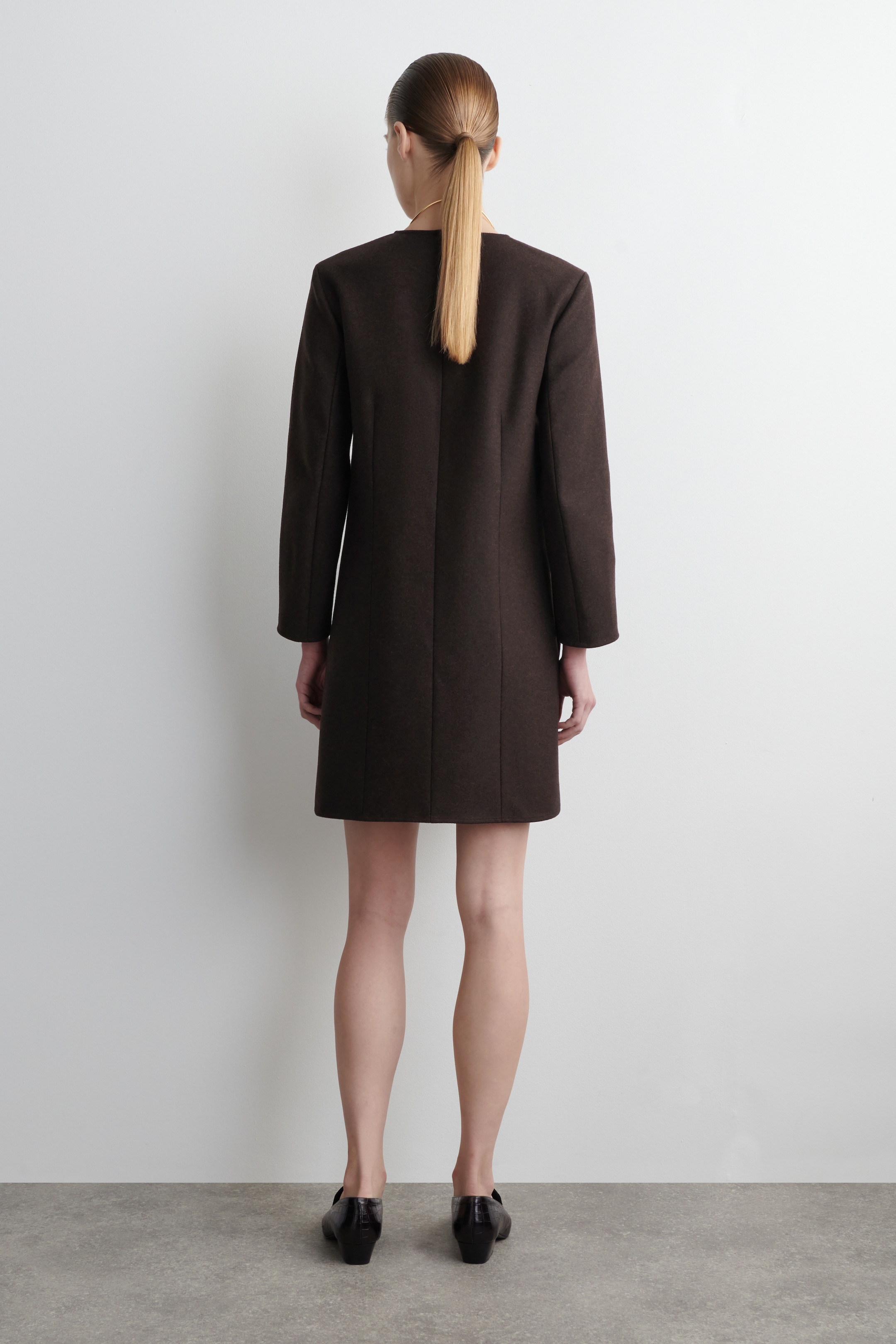 Ver imagen más grande: Se muestra un vestido marrón oscuro desde atrás, con un diseño recto y corto que termina por encima de las rodillas. Está diseñado con mangas largas y un escote sin cuello.