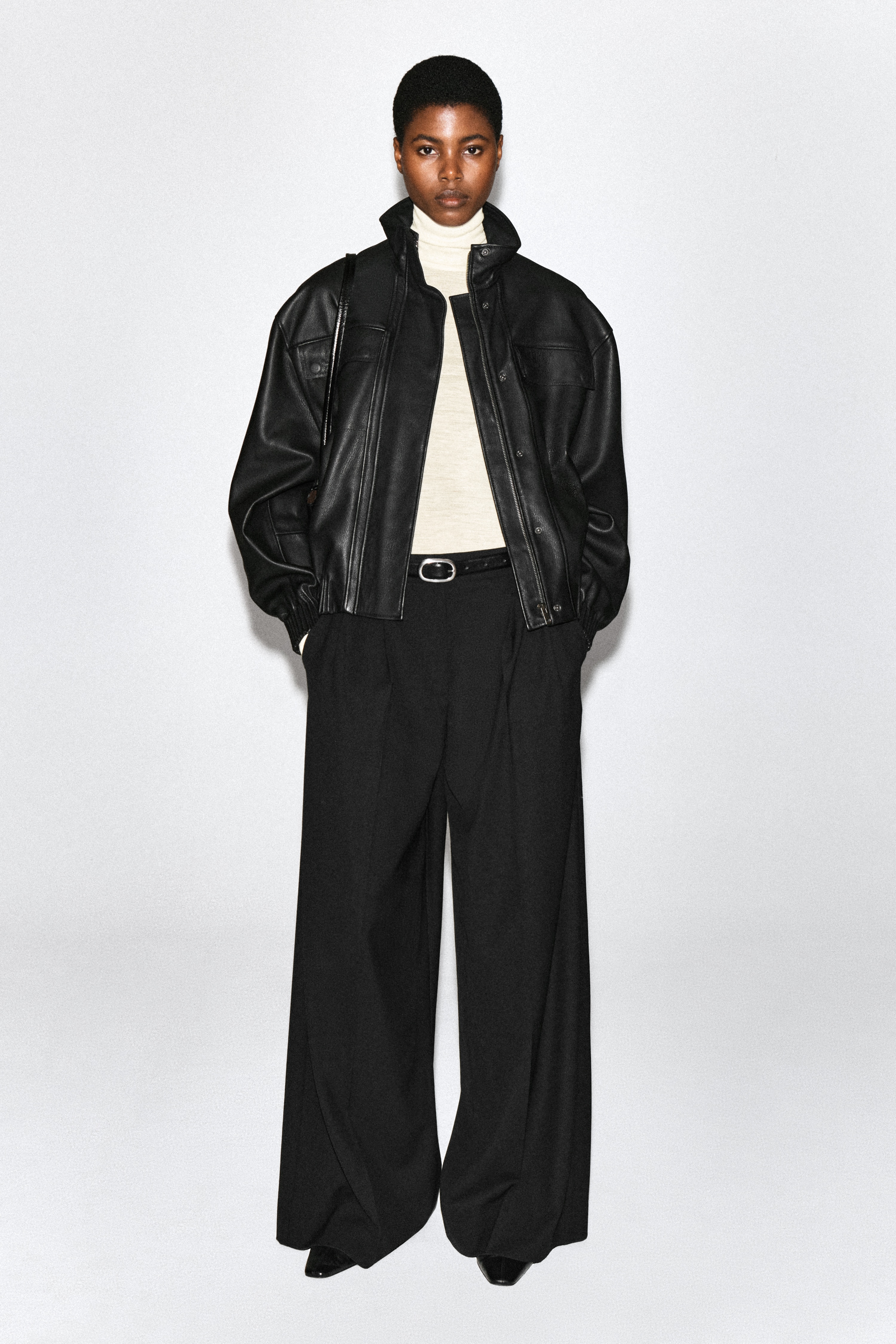 Pantalones anchos plisados en negro mostrados desde una vista frontal, con cintura estructurada, pliegues delanteros profundos y bolsillos laterales. Las perneras caen en una silueta de pierna ancha con marcadas rayas planchadas que recorren el centro.
