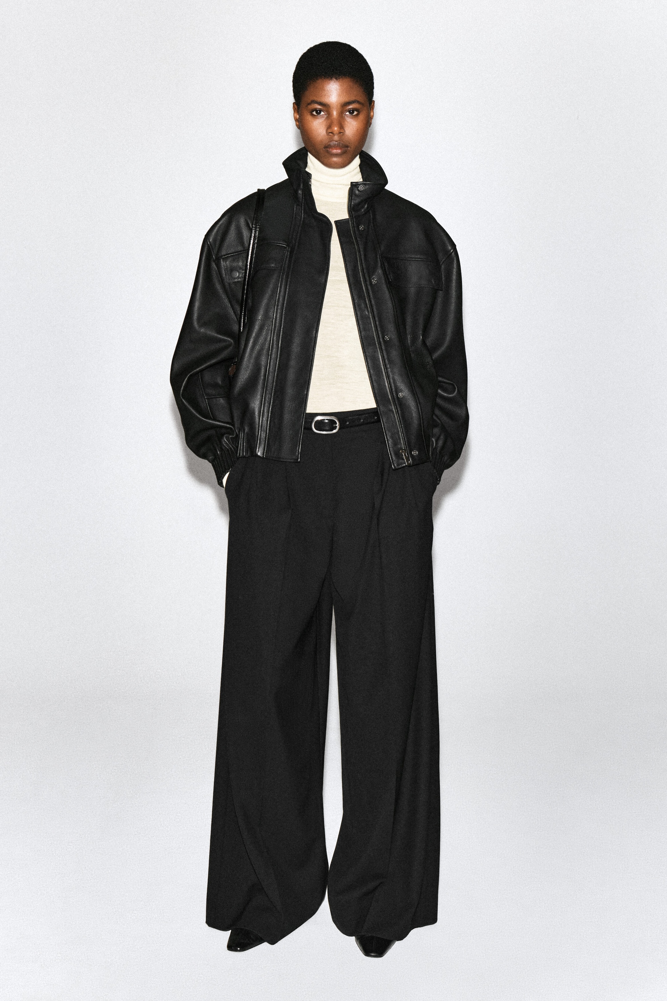 Pleated wide-leg pants in black are being worn by the model, who is standing with hands tucked into the side pockets. The high-waisted, relaxed-fit pants are styled with a thin black belt, a cream turtleneck top, and an oversized black leather jacket. Pleated wide-leg pants in black are being worn by the model, who is standing with hands tucked into the side pockets. The high-waisted, relaxed-fit pants are styled with a thin black belt, a cream turtleneck top, and an oversized black leather jacket.