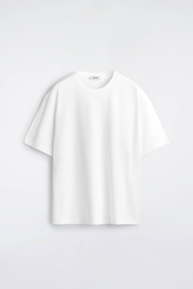 A white T-shirt is shown from the front, featuring a relaxed fit, a ribbed crew neck, and short sleeves. A white label with "COS" in black lettering is visible inside the neckline. - 1