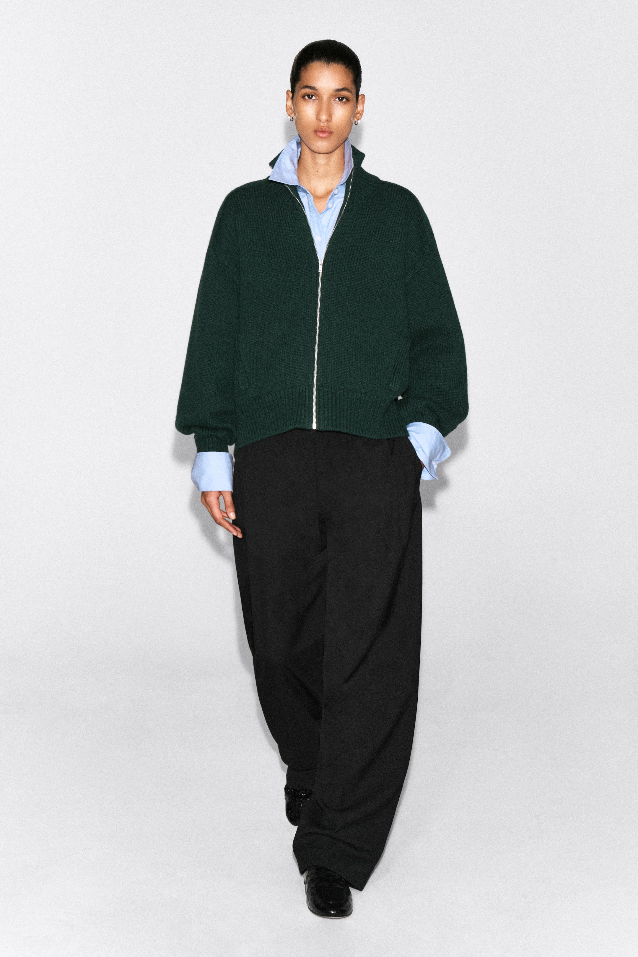 Visualizza immagine più grande: Una modella indossa una giacca zip-up in lana verde scuro con collo a imbuto arrotondato, ripresa frontalmente, che mette in risalto il suo fit rilassato e la texture a maglia a coste. La giacca presenta una cerniera frontale color argento parzialmente aperta per rivelare una camicia azzurra a colletto sovrapposta, oltre a dettagli a coste su polsini e orlo.