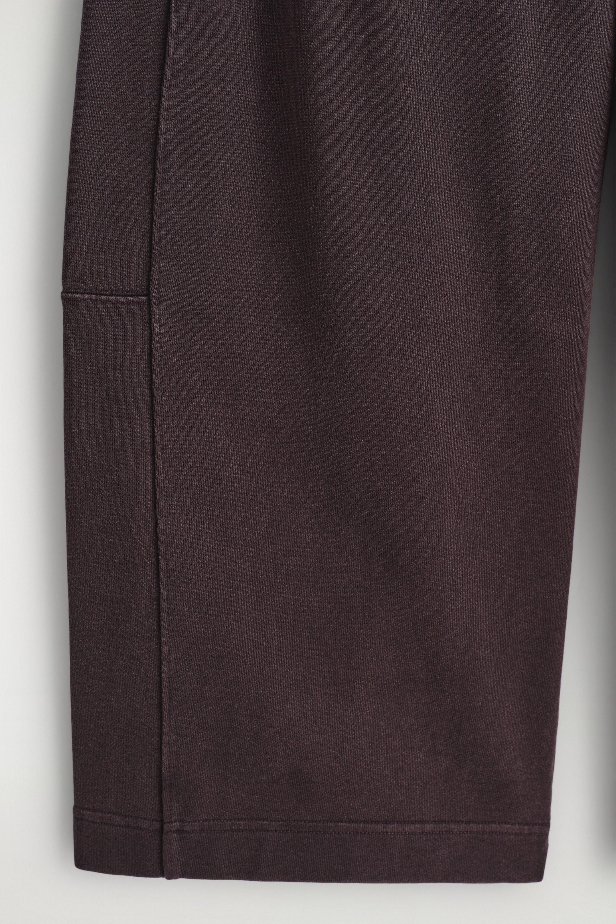 View larger image: A dark brown garment leg is being presented straight-on, displaying its subtle fabric texture, a prominent vertical seam running down the left side, and a cleanly finished hem with a stitched panel detail above it.