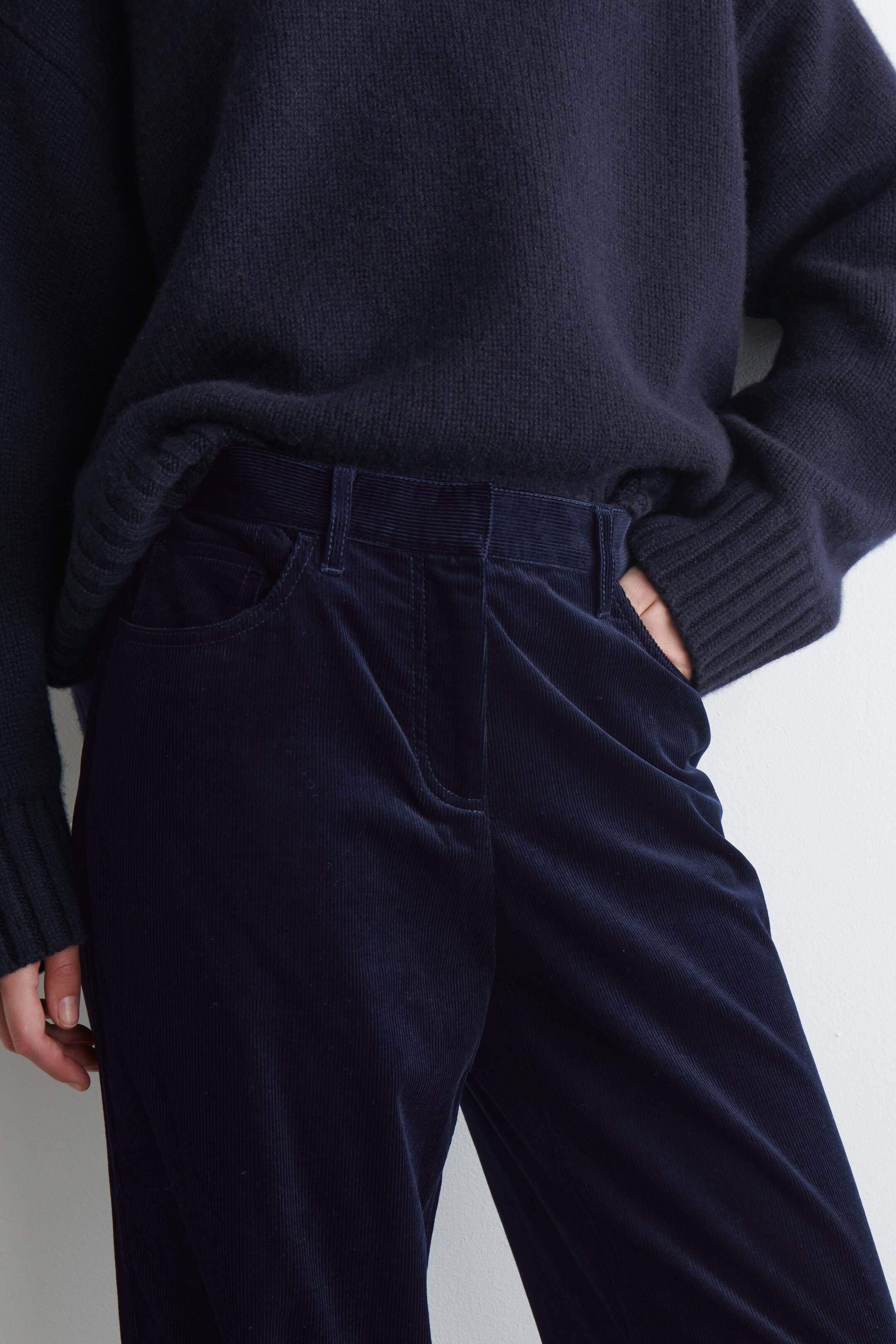 Agrandir l'image: Un pantalon en velours côtelé bleu foncé est porté, avec des côtes visibles, des passants de ceinture et des poches avant en biais, montré sous un angle légèrement surélevé, se concentrant sur la taille et la hanche. Un pull en tricot bleu foncé avec des poignets côtelés est superposé sur le pantalon, avec une main reposant dans une poche du pantalon.