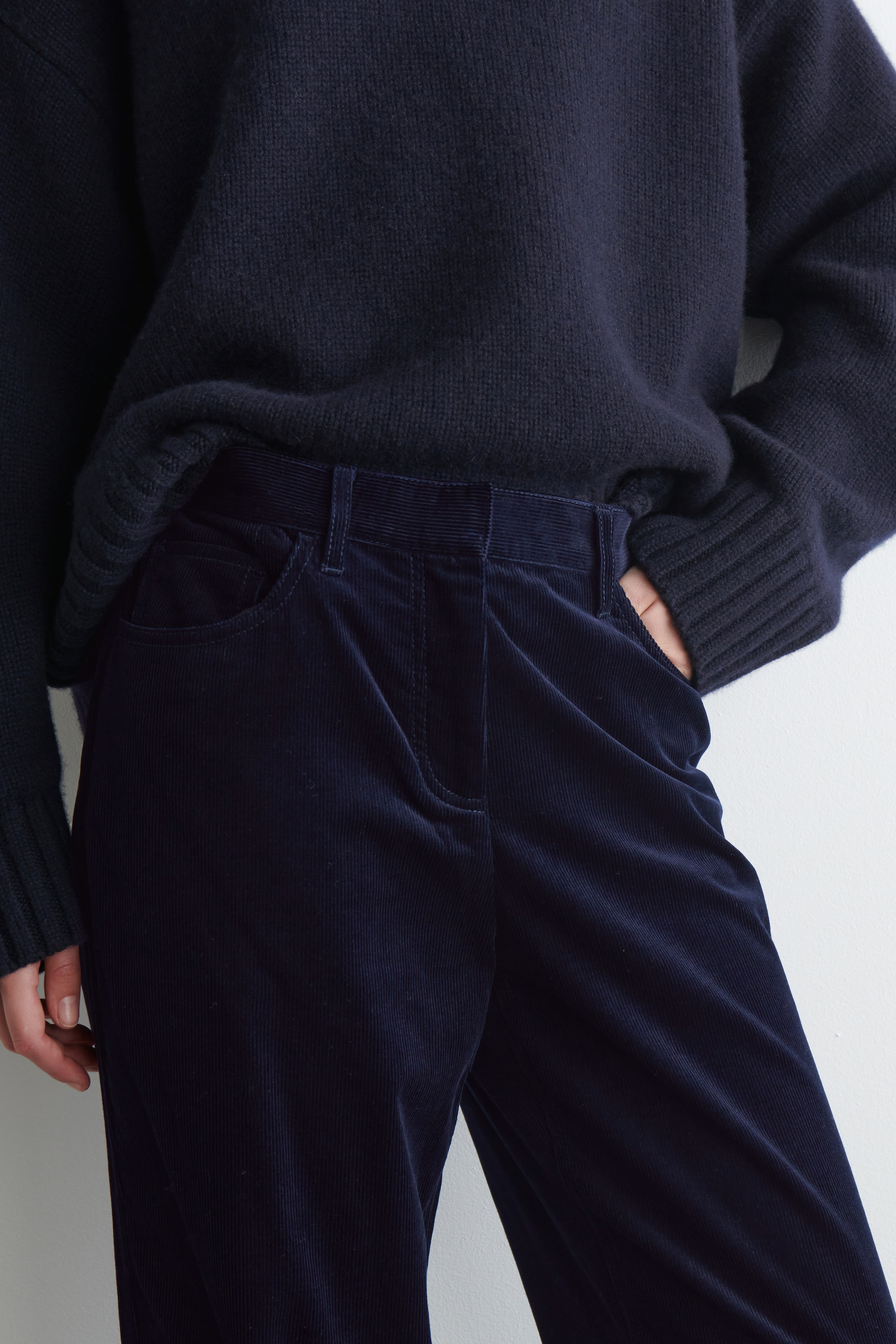 Größeres Bild ansehen: Eine dunkelblaue Cordhose wird getragen, mit sichtbaren Rippen, Gürtelschlaufen und vorderen Schlitztaschen, aus einem leicht erhöhten Winkel von vorne gezeigt, der sich auf die Taille und die Hüfte konzentriert. Ein dunkelblauer Strickpullover mit gerippten Bündchen wird über die Hose geschichtet, wobei eine Hand in einer Hosentasche ruht.