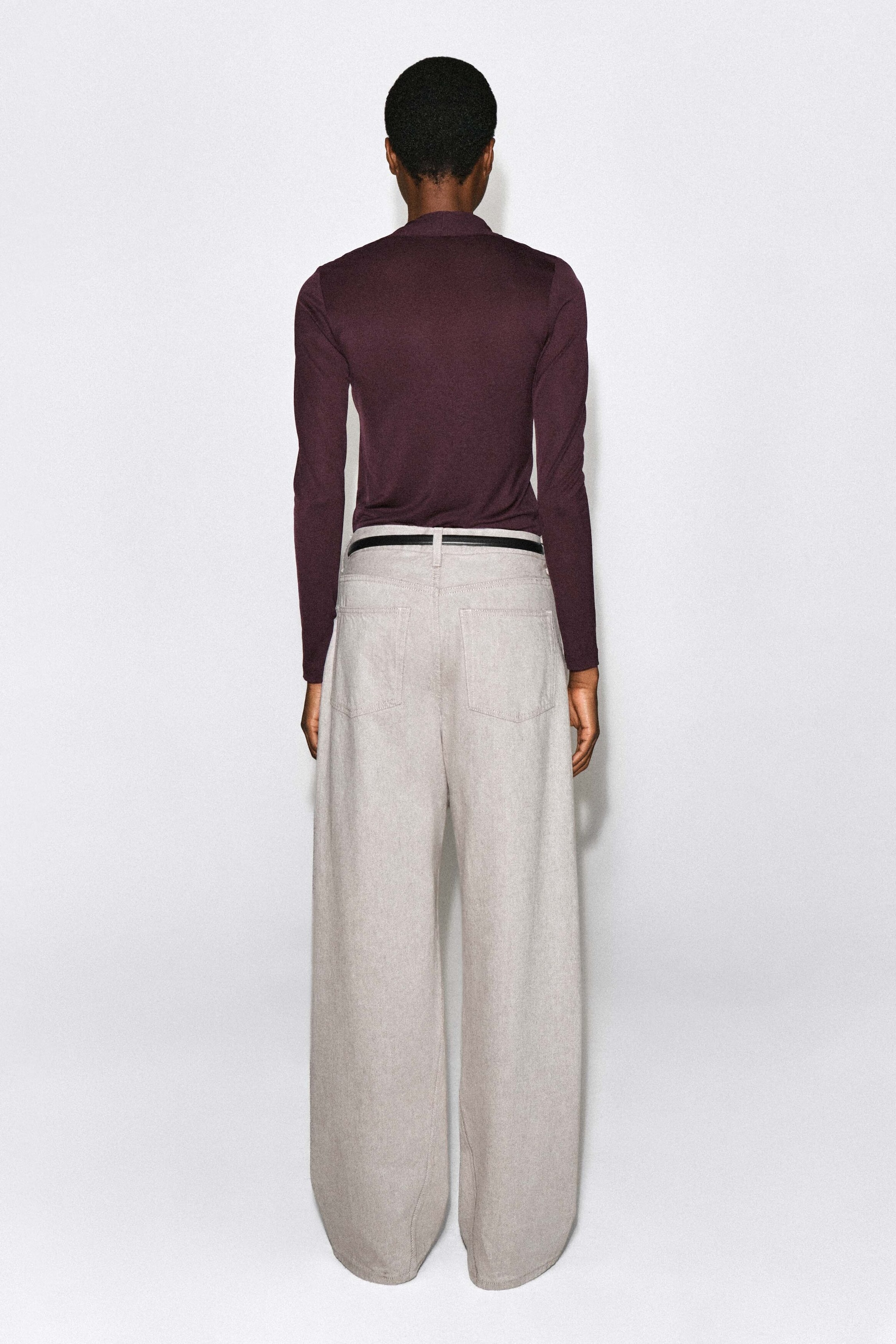 A dark purple, long-sleeved top is worn by the model, viewed from the back to show the high collar detail. The slim-fit top is tucked into light gray, wide-leg trousers styled with a thin black belt. A dark purple, long-sleeved top is worn by the model, viewed from the back to show the high collar detail. The slim-fit top is tucked into light gray, wide-leg trousers styled with a thin black belt.
