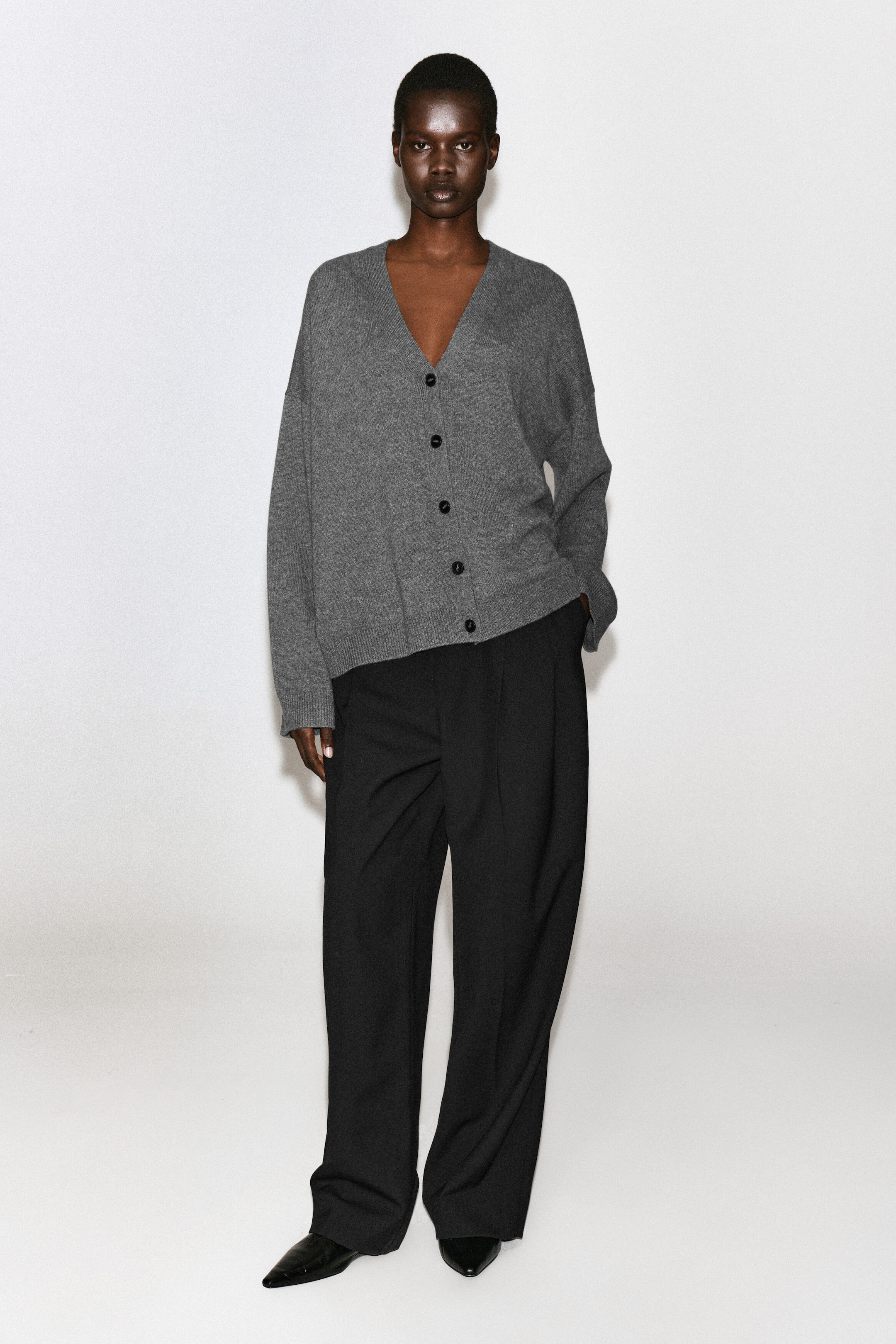 Un cardigan in cashmere grigio, caratterizzato da uno scollo a V profondo, chiusure frontali con bottoni scuri e bordi a coste, indossato dalla modella. Il cardigan è abbinato a pantaloni neri a gamba ampia e scarpe nere con punta affusolata. Un cardigan in cashmere grigio, caratterizzato da uno scollo a V profondo, chiusure frontali con bottoni scuri e bordi a coste, indossato dalla modella. Il cardigan è abbinato a pantaloni neri a gamba ampia e scarpe nere con punta affusolata.