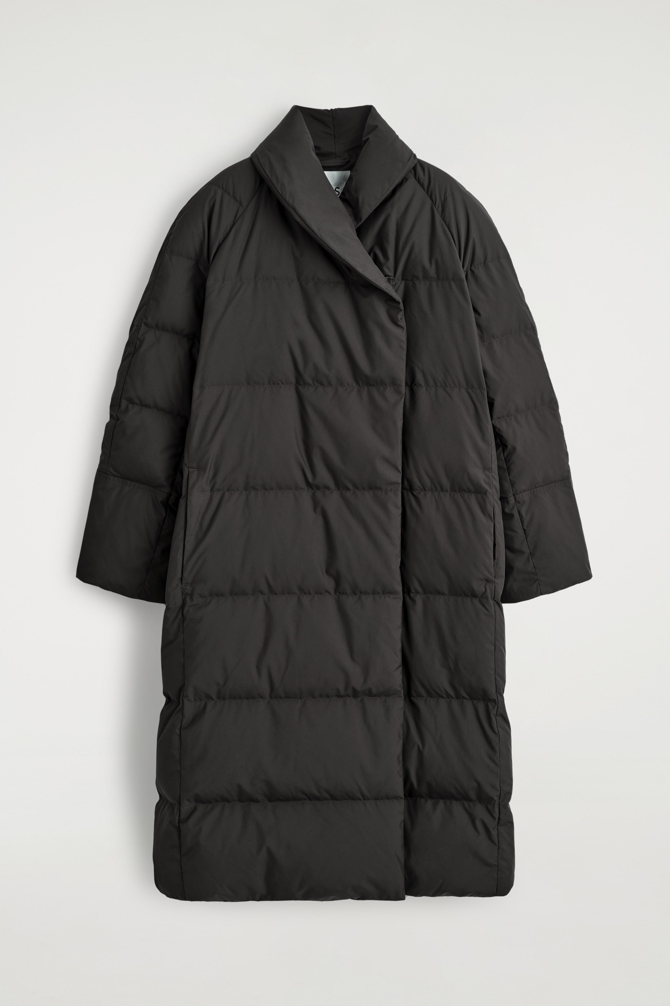 View larger image: A dark gray longline puffer coat is presented FRONT, featuring a wide shawl collar, horizontal quilting, and raglan sleeves. The front is showing an overlapping panel secured by a single closure point near the collar, and subtle vertical side pockets are visible.