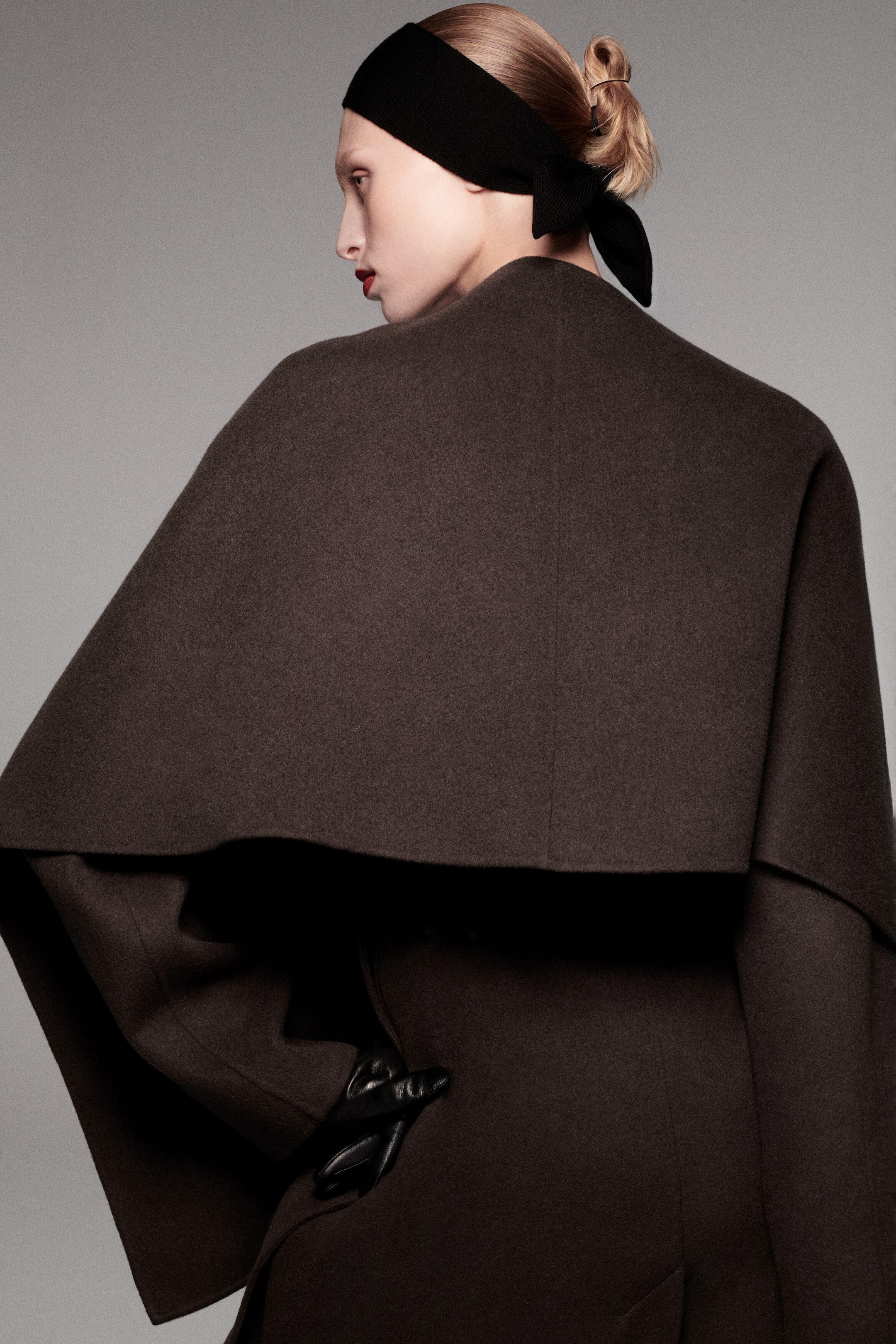 A double-faced wool-blend cape in dark brown is shown from the back, featuring a structured capelet-style overlay that drapes over the shoulders and upper back. A hand wearing a black leather glove rests on the hip, highlighting the open, layered design of the cape. A double-faced wool-blend cape in dark brown is shown from the back, featuring a structured capelet-style overlay that drapes over the shoulders and upper back. A hand wearing a black leather glove rests on the hip, highlighting the open, layered design of the cape.