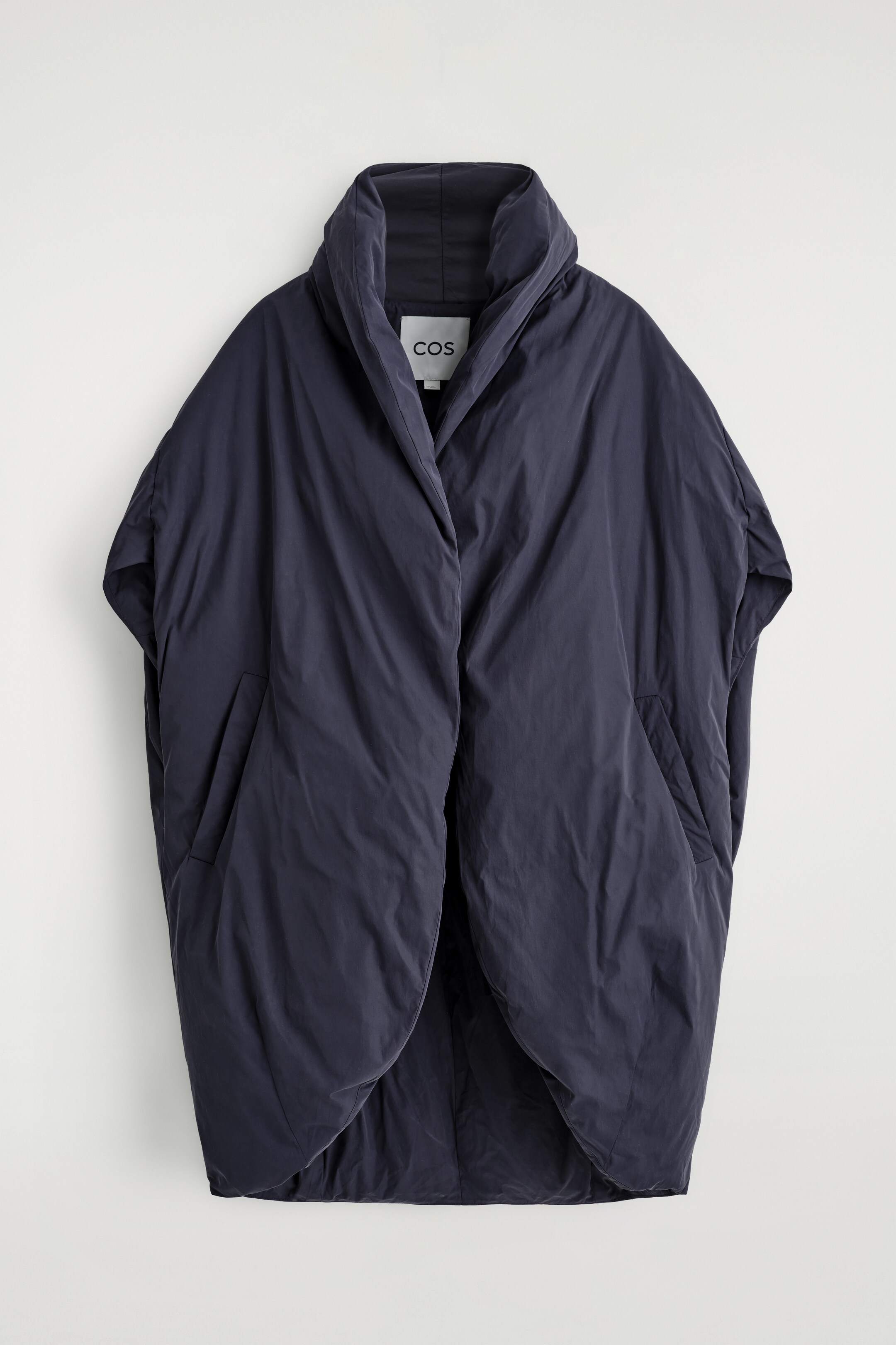 Ver imagen más grande: Se presenta un abrigo estilo poncho acolchado de color azul oscuro desde el frente, que cuenta con un cuello ancho y drapeado, un frente abierto con paneles superpuestos y dos bolsillos inclinados con ranura, con una etiqueta 'COS' visible en el cuello interior.