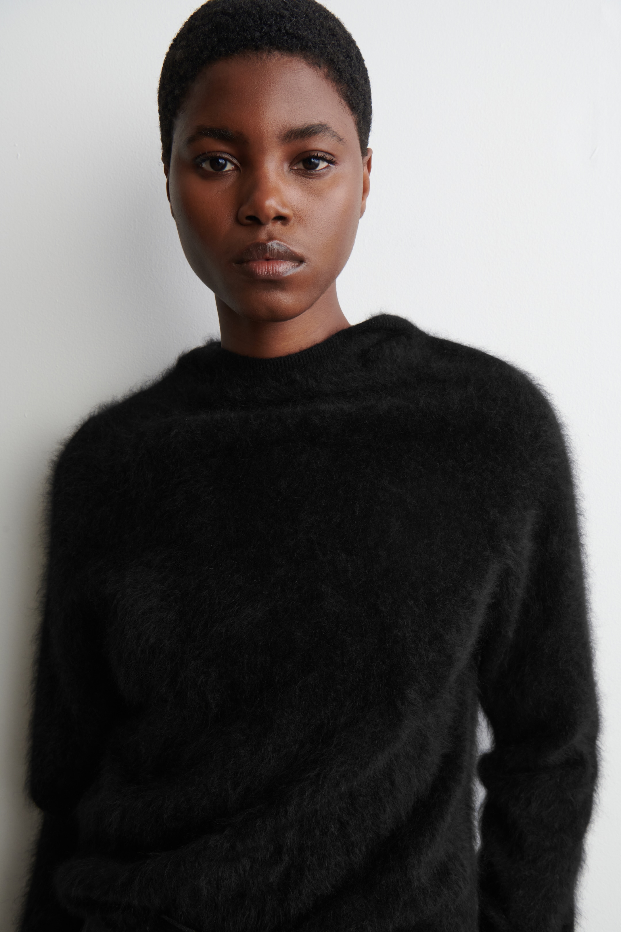 View larger image: A black, fuzzy sweater is being presented, seen from the front and angled slightly to the left, revealing its relaxed fit. It is featuring a round crew neck and a prominent, soft shaggy texture throughout.