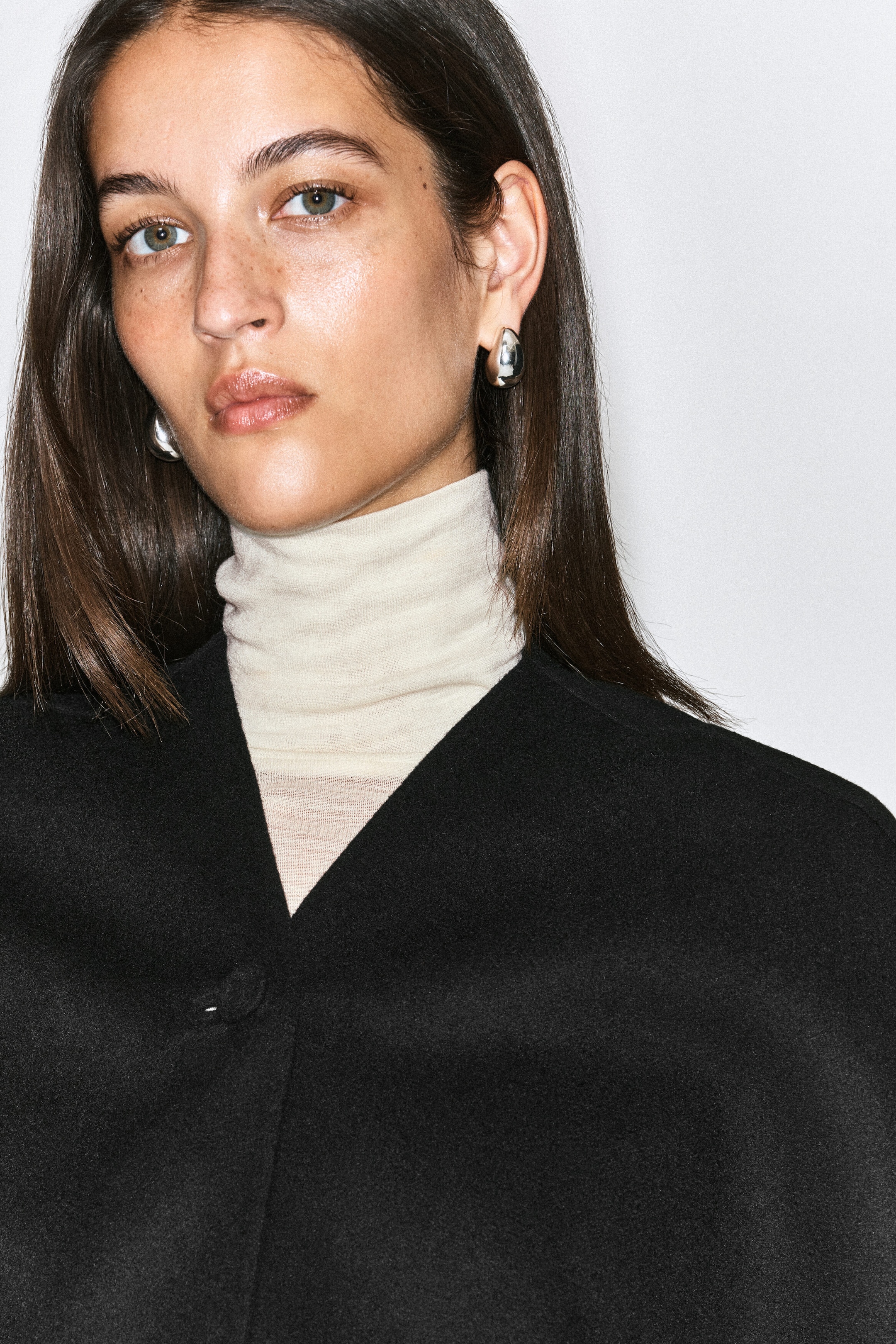 A black double-faced wool-blend cape featuring a deep V-neckline and a single-button fastening is worn by the model. The cape is layered over a cream high-neck top, highlighting its minimalist, structured design. A black double-faced wool-blend cape featuring a deep V-neckline and a single-button fastening is worn by the model. The cape is layered over a cream high-neck top, highlighting its minimalist, structured design.