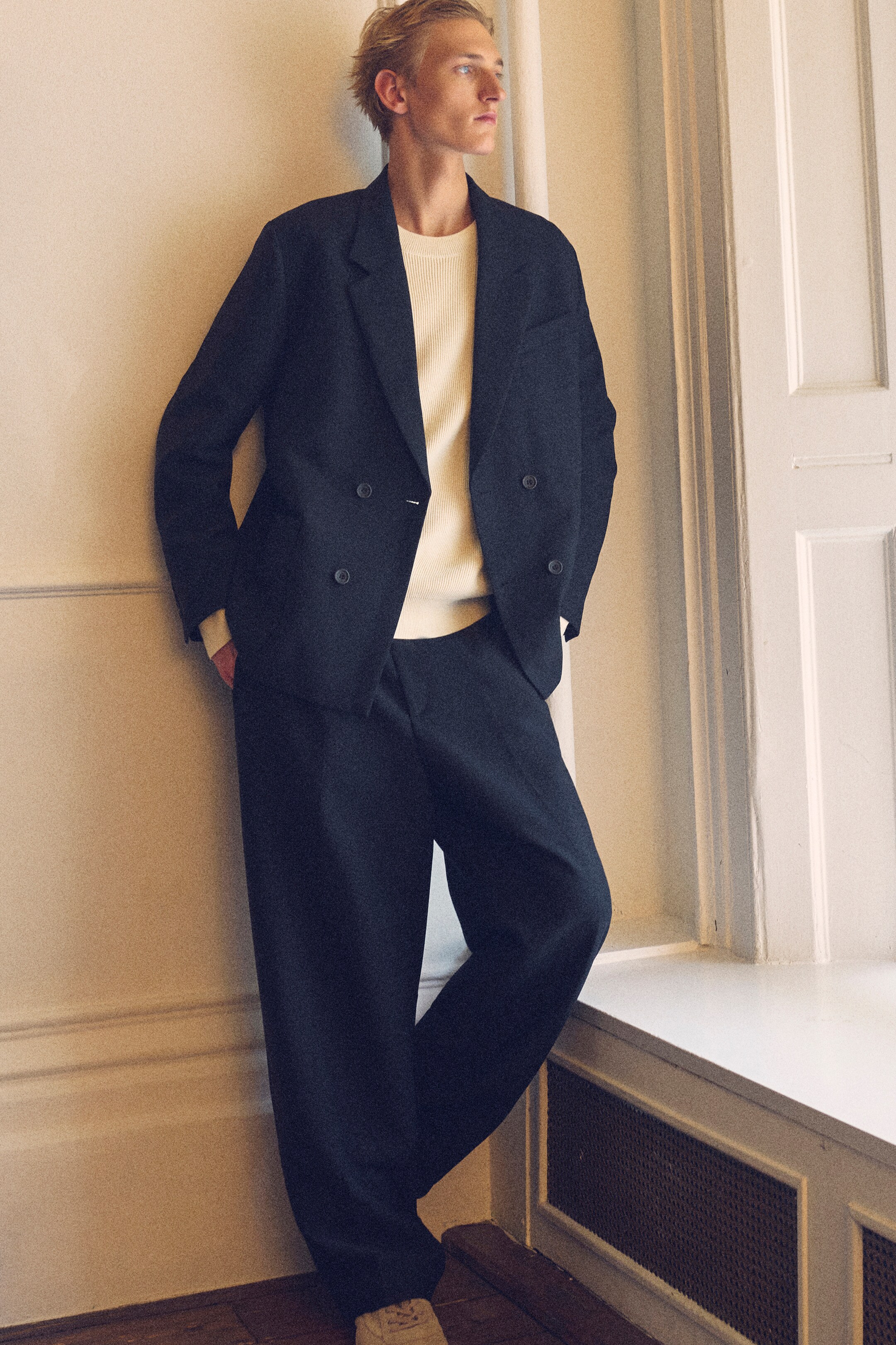 Größeres Bild anzeigen: Ein dunkler, zweireihiger Blazer mit einer entspannten, unstrukturierten Passform und Kerbrevers wird von vorne präsentiert und zeigt zwei Knopfreihen. Er wird mit dunklen, gerade geschnittenen Hosen kombiniert, die einen mühelosen Fall aufweisen, und hellen Schnürtrainern.