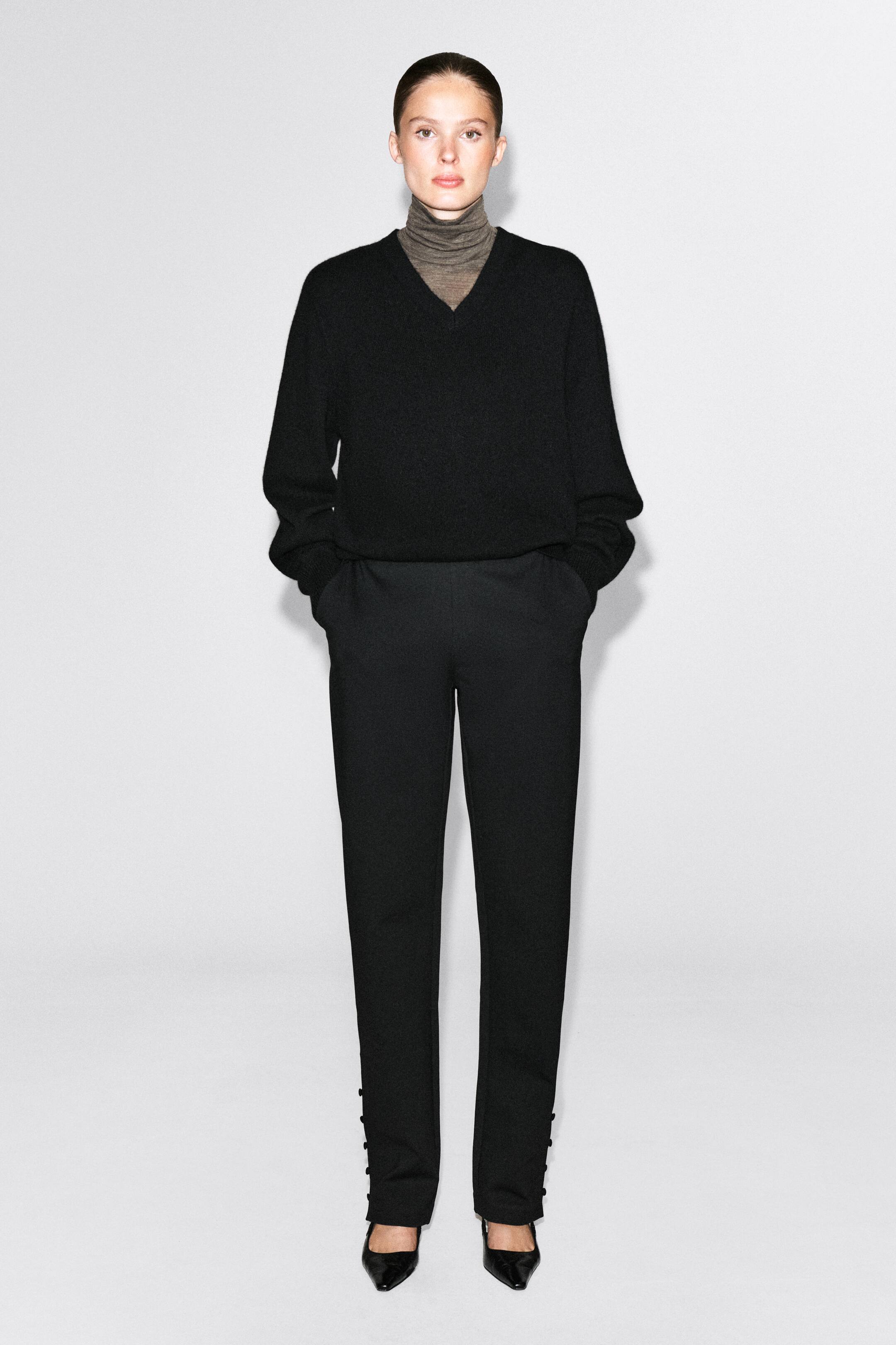 Ver imagen más grande: Unos pantalones negros de corte slim con detalles de botones forrados en tejido en el exterior de los bajos del tobillo los lleva la modelo, que posa con las manos en los bolsillos laterales. Los pantalones se combinan con un jersey negro de cuello en V superpuesto sobre un cuello alto gris y se completan con unos zapatos de tacón negros de punta fina.