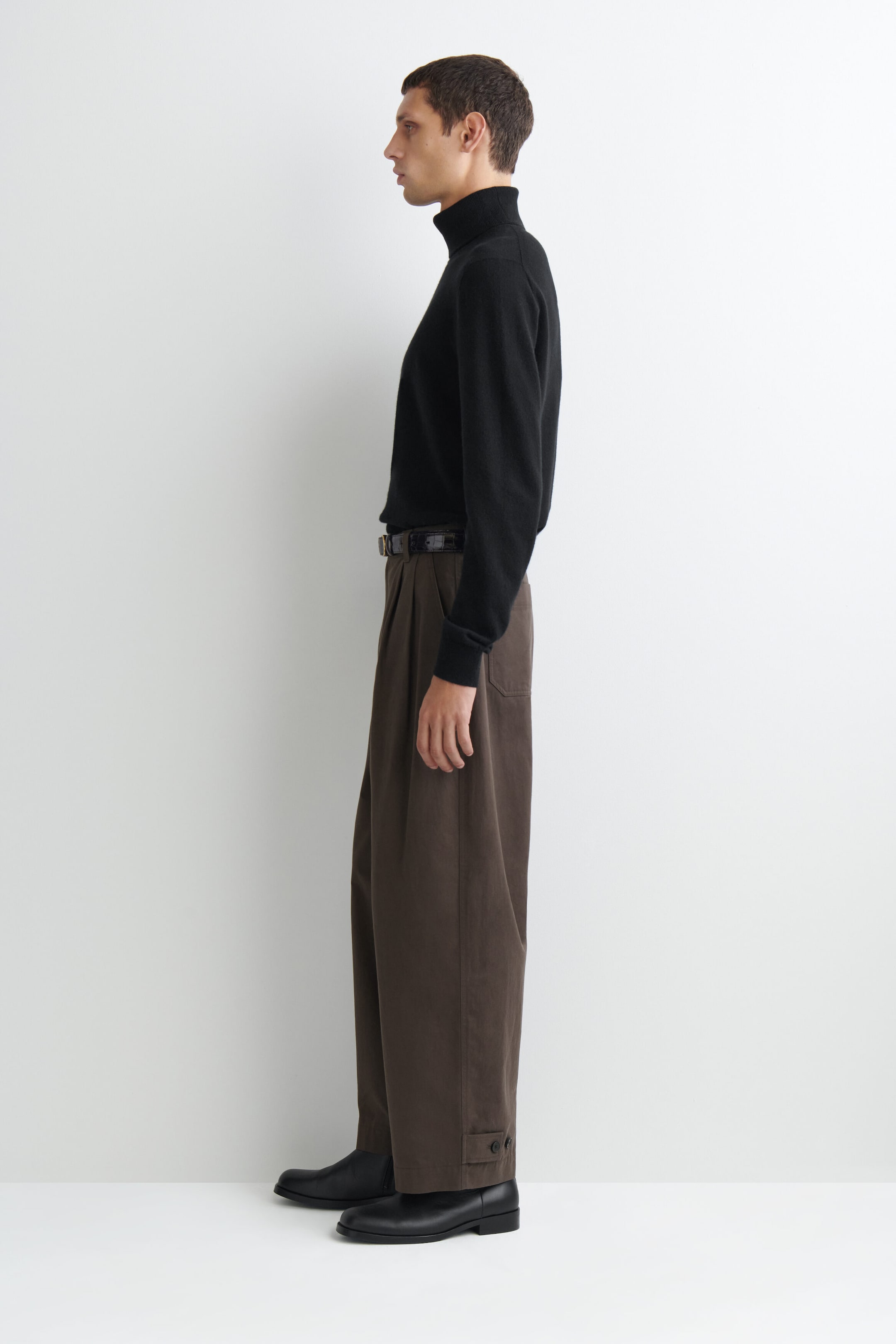 A black knit turtleneck sweater and dark brown wide-leg trousers are shown worn facing left to highlight their relaxed silhouette. The long-sleeve sweater features a ribbed collar and cuffs, while the pleated trousers feature a high-rise waist with a black belt and adjustable button tabs at the hems. A black knit turtleneck sweater and dark brown wide-leg trousers are shown worn facing left to highlight their relaxed silhouette. The long-sleeve sweater features a ribbed collar and cuffs, while the pleated trousers feature a high-rise waist with a black belt and adjustable button tabs at the hems.