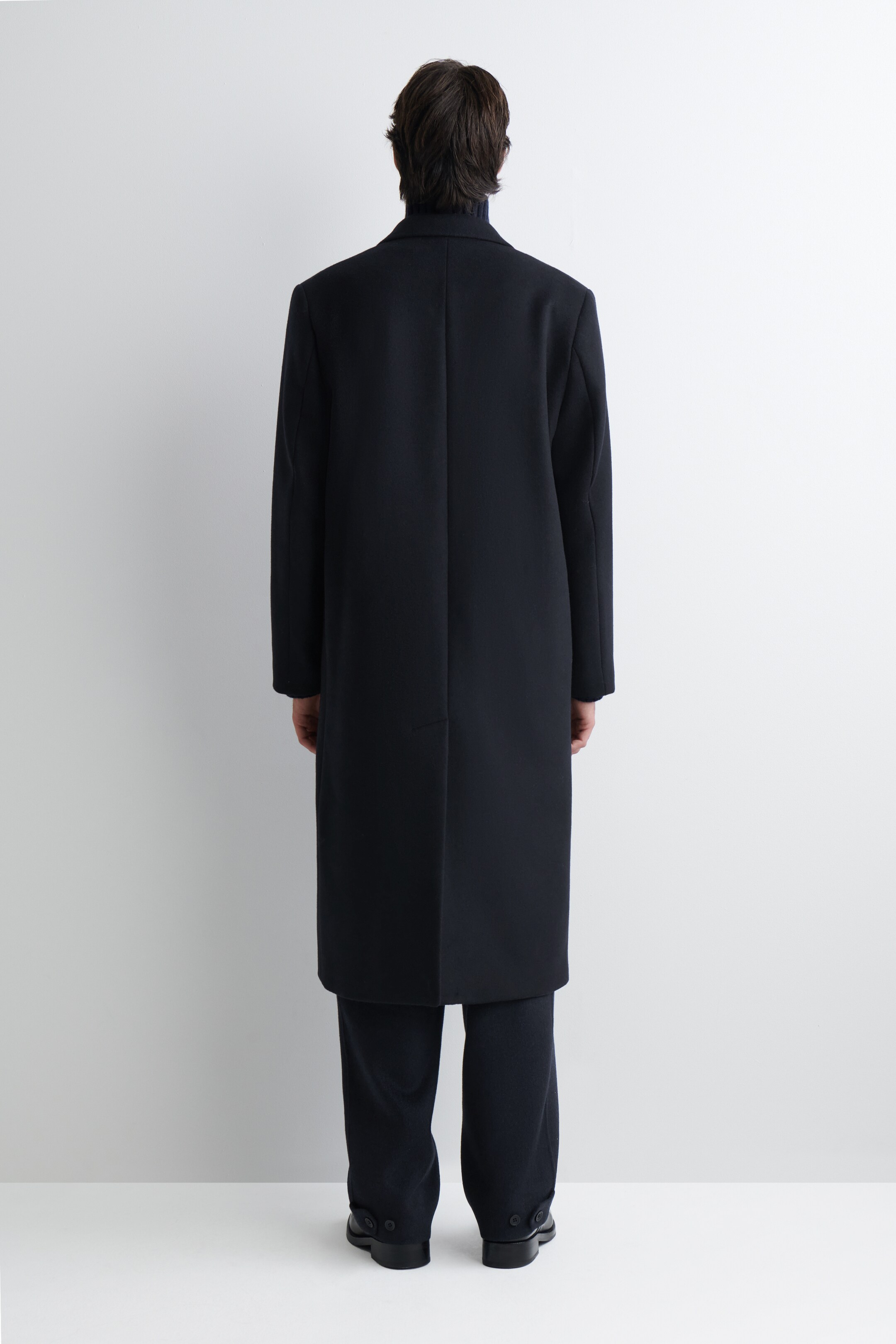 View larger image: A long, dark coat is being presented from the back view, featuring a straight silhouette that extends to the ankles. A visible center back seam is running down the length of the coat, which is being worn over a dark turtleneck and trousers.