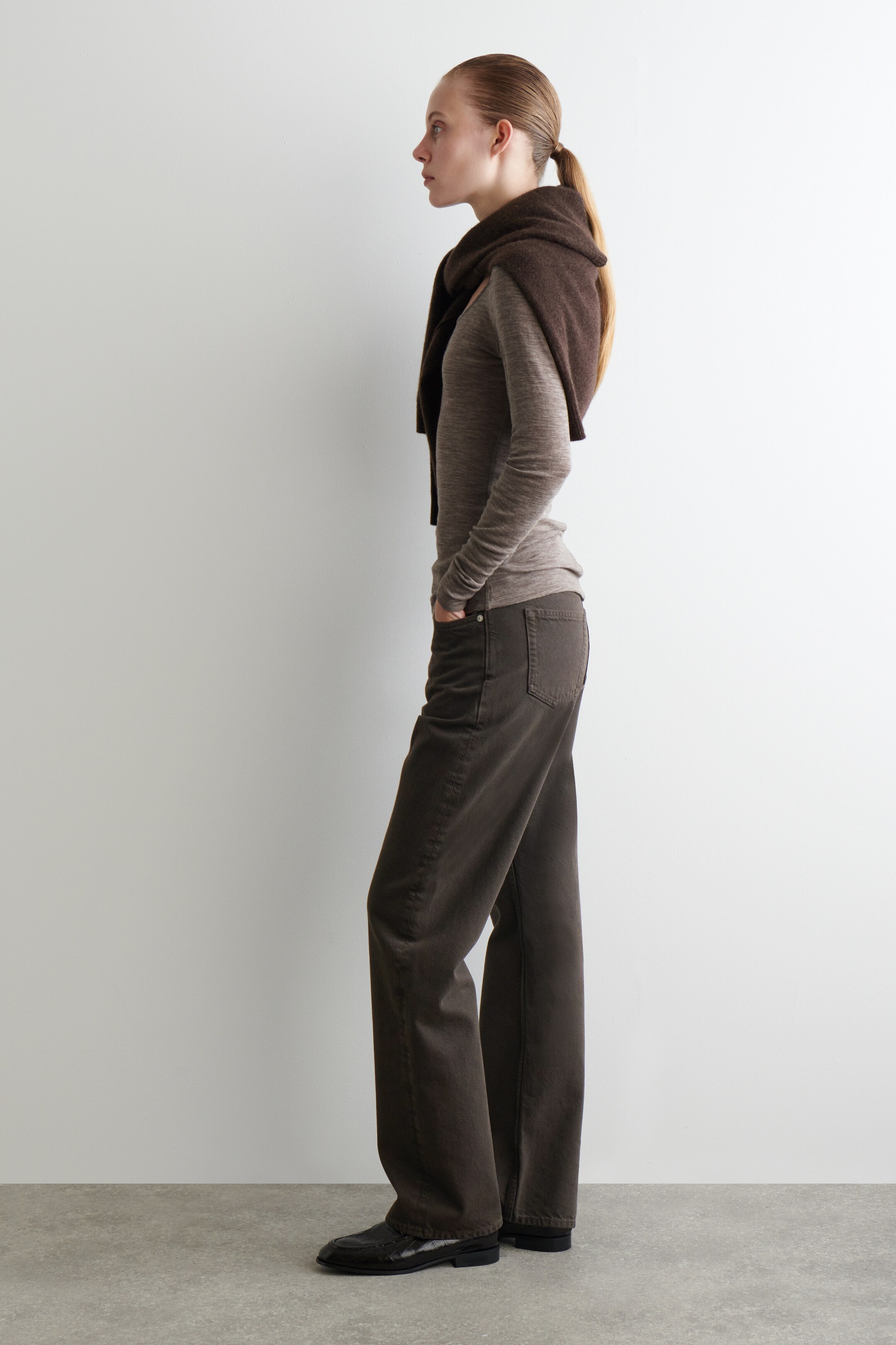 Vezi imaginea mai mare: Un top tricotat cu mâneci lungi, de culoare taupe deschis, este purtat dintr-un unghi lateral, stratificat cu un pulover tricotat de culoare maro închis, drapat peste umeri. Pantaloni maro închis, cu croială lejeră și picioare largi, cu un buzunar din spate vizibil, sunt purtați împreună cu mocasini negri cu vârf pătrat.