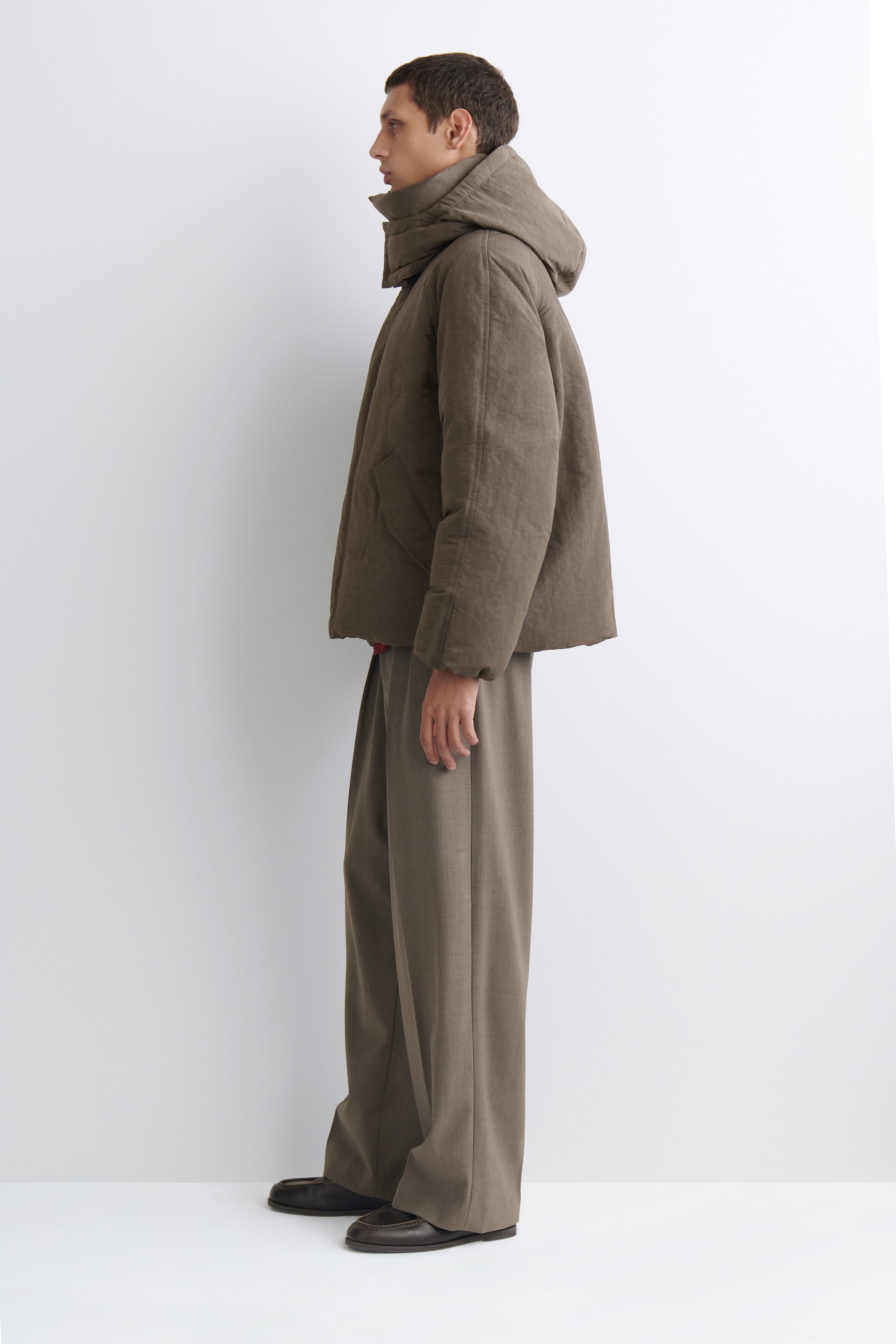 The oversized nylon-linen hooded down puffer jacket is shown facing left, highlighting its boxy, voluminous silhouette and textured taupe-brown fabric. It features a high-standing padded collar, a large hood, and subtle welt pockets, while draping over wide-leg trousers to emphasize its relaxed fit. The oversized nylon-linen hooded down puffer jacket is shown facing left, highlighting its boxy, voluminous silhouette and textured taupe-brown fabric. It features a high-standing padded collar, a large hood, and subtle welt pockets, while draping over wide-leg trousers to emphasize its relaxed fit.