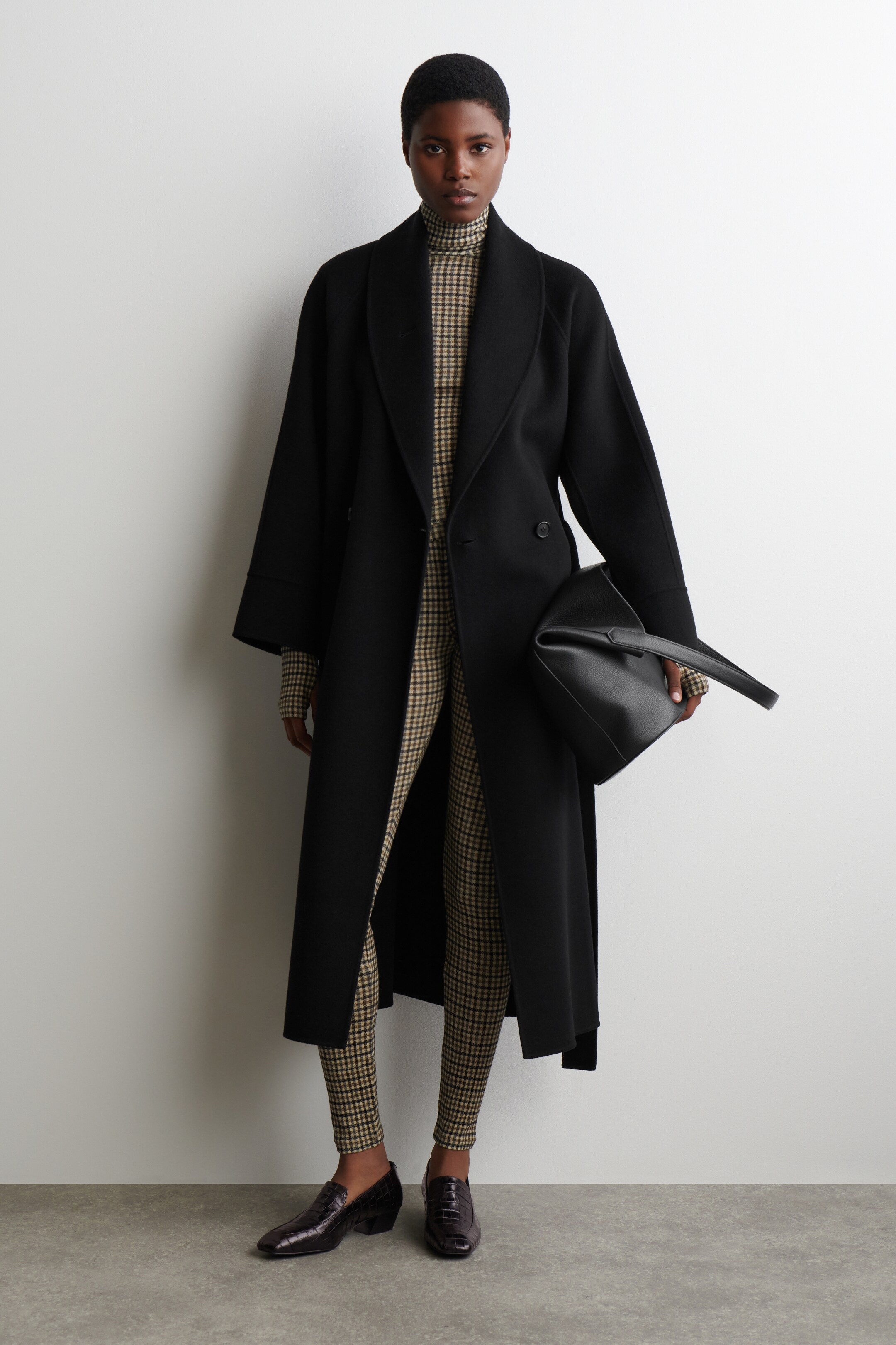 Größeres Bild anzeigen: Ein langer schwarzer Mantel wird präsentiert, der einen breiten, weichen Schalkragen und eine entspannte, offene Silhouette aufweist. Er wird über einem braun-schwarz karierten Rollkragenpullover und passenden Hosen getragen, wobei eine schwarze strukturierte Handtasche und dunkle, strukturierte Loafer den Look vervollständigen.
