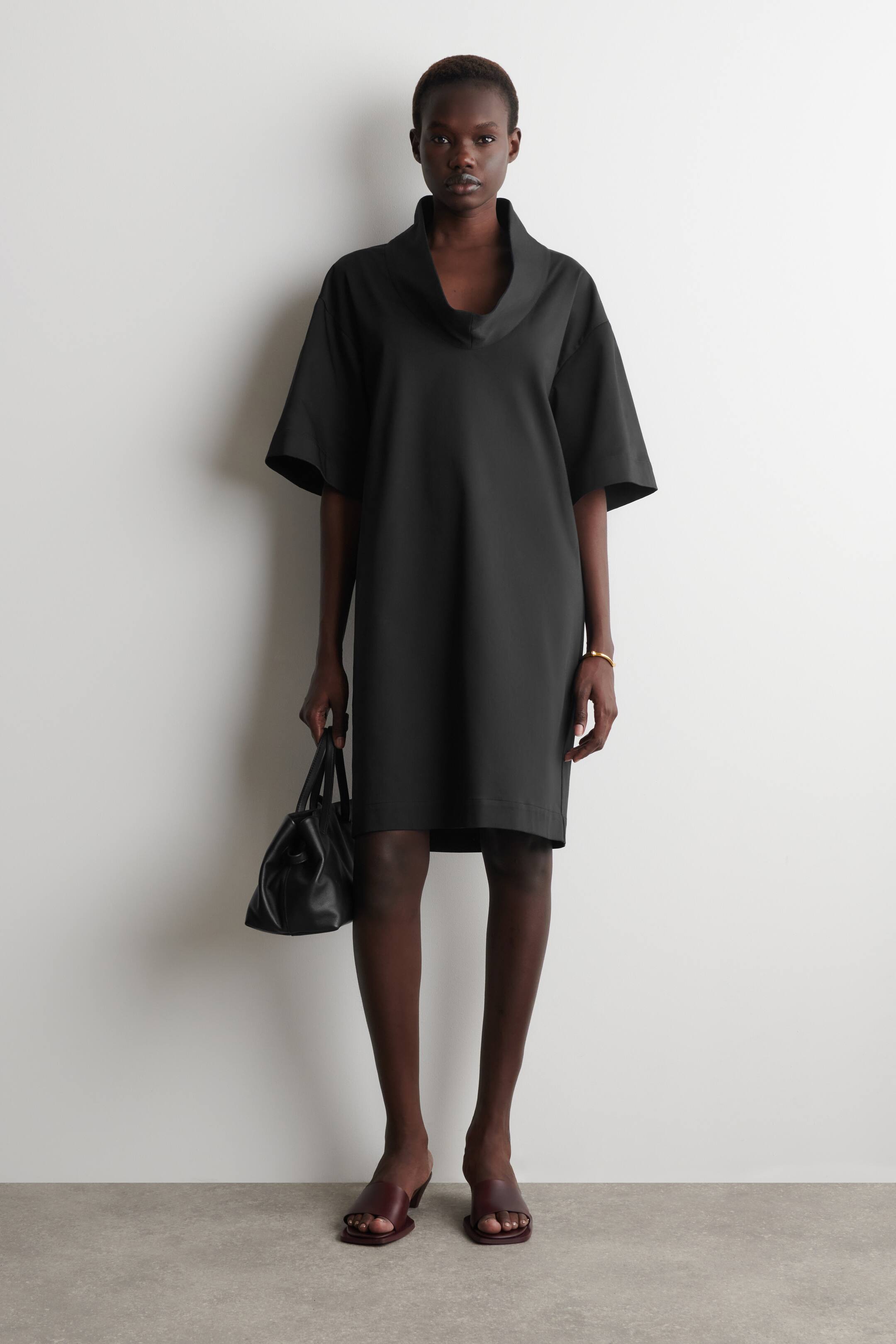 Agrandir l'image: La robe noire est présentée de face, avec un col haut drapé et des manches courtes, larges et tombantes. Elle offre une silhouette ample et droite, tombant au-dessus du genou.