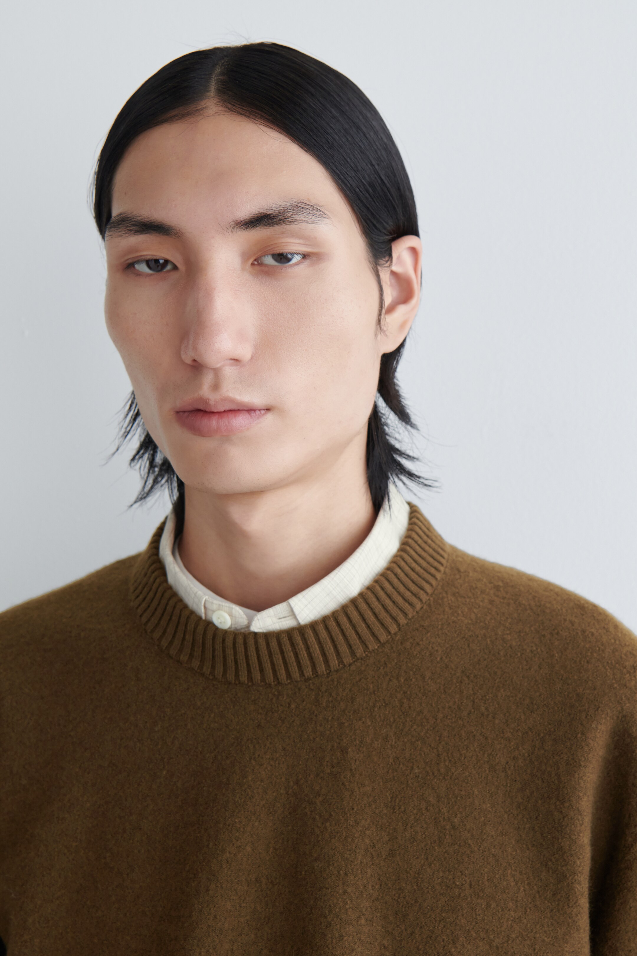 Agrandir l'image: Un pull à col rond brun terreux est présenté, avec un col côtelé proéminent et un tricot doux, légèrement texturé. Une chemise à col clair, subtilement à motifs, est portée en dessous, avec son bouton supérieur fermé et le col visible.