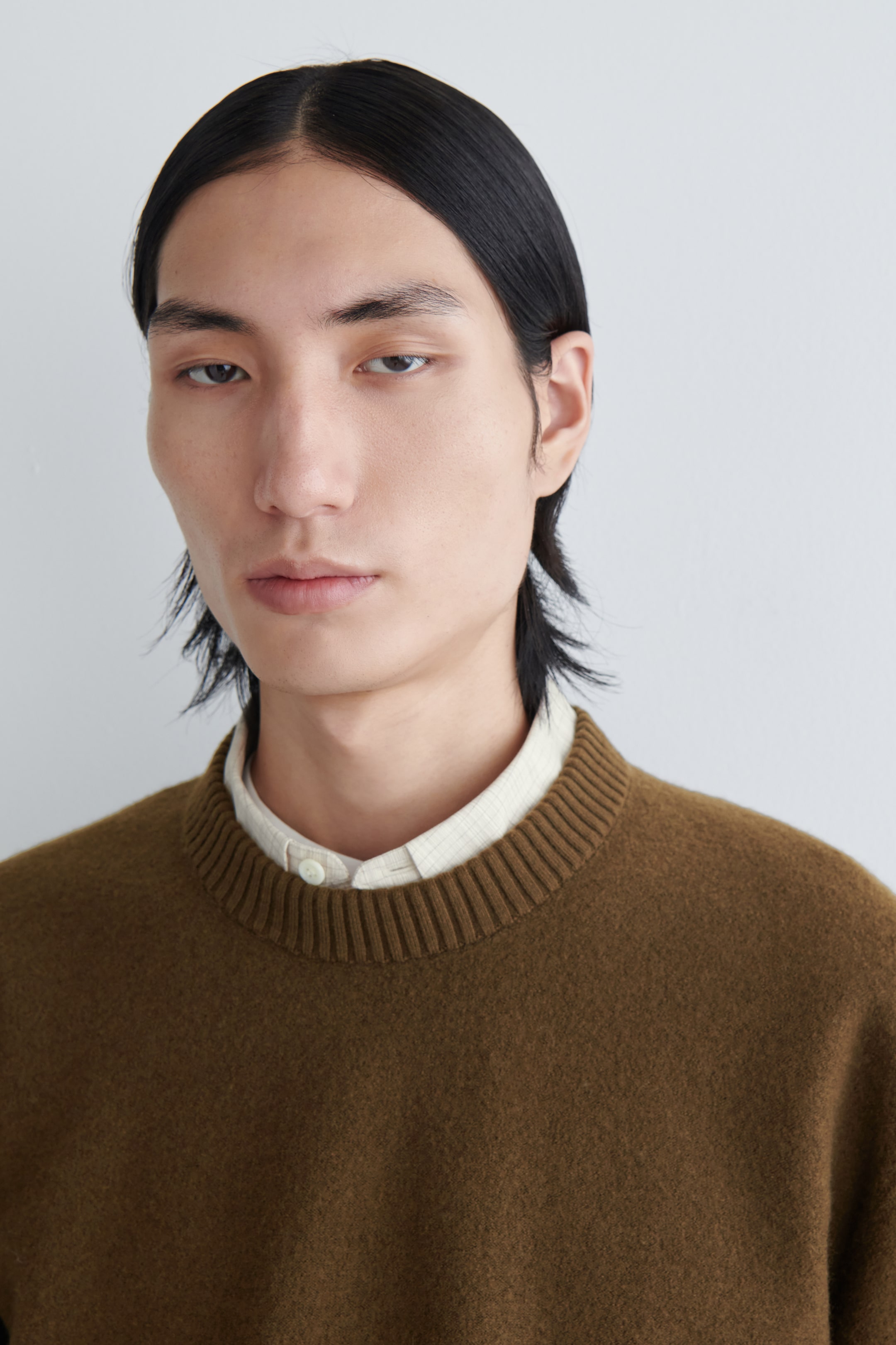 View larger image: An earthy brown crew neck sweater is being presented, featuring a prominent ribbed collar and a soft, slightly textured knit. A light-colored, subtly patterned collared shirt is being worn underneath, with its top button fastened and collar visible.