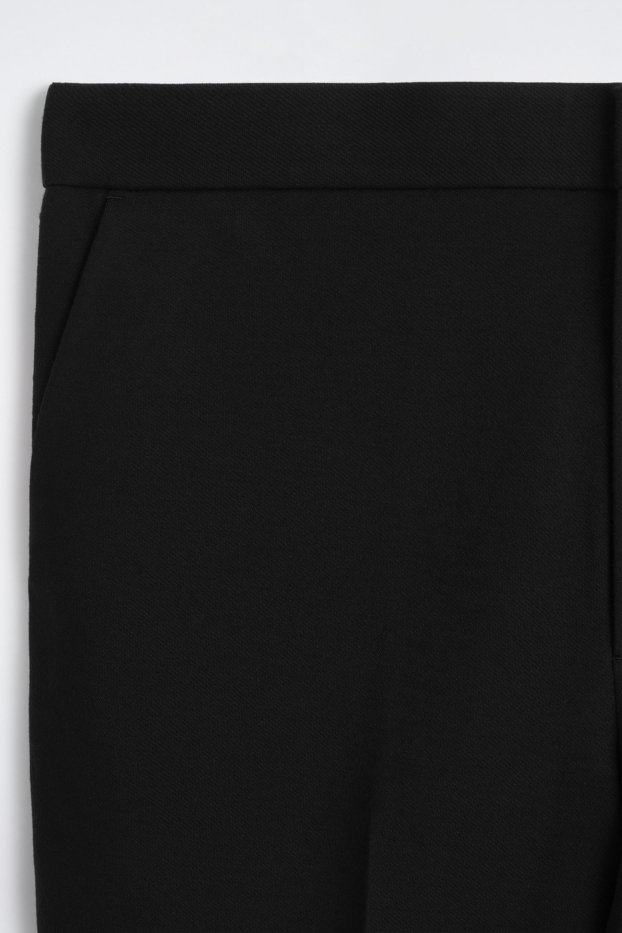 Black trousers feature a clean-cut waistband and a subtle side slant pocket in a close-up front view. The fabric shows a fine, textured diagonal weave. Black trousers feature a clean-cut waistband and a subtle side slant pocket in a close-up front view. The fabric shows a fine, textured diagonal weave.