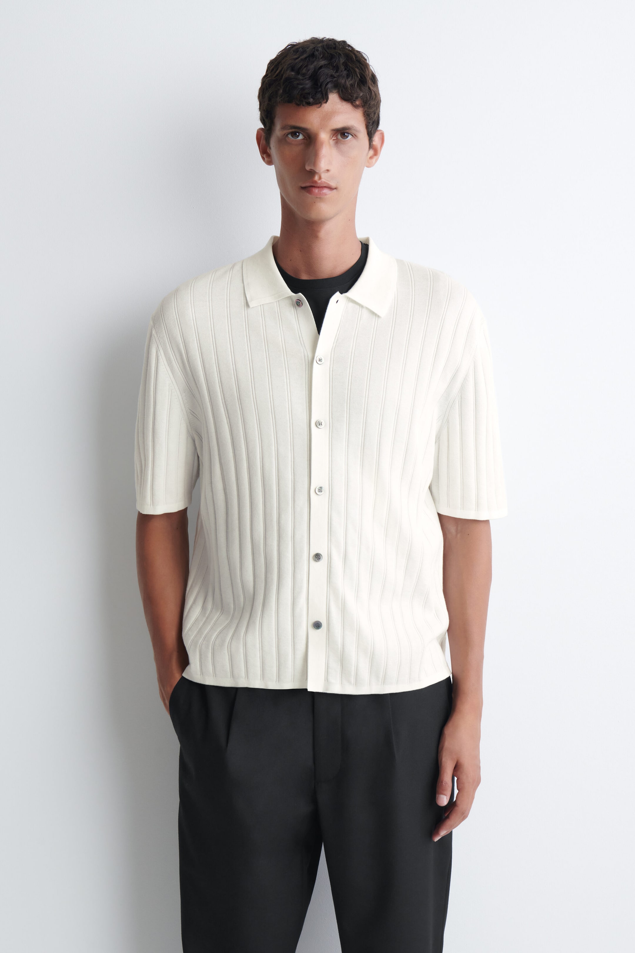 Ein Shirt aus gerippter Baumwolle in Off-White wird vom Model getragen und verfügt über eine vertikale Rippstruktur, eine durchgehende Knopfleiste und einen klassischen Kragen. Das kurzärmelige Shirt wird über einem schwarzen T-Shirt getragen und mit einer schwarzen Hose kombiniert. Ein Shirt aus gerippter Baumwolle in Off-White wird vom Model getragen und verfügt über eine vertikale Rippstruktur, eine durchgehende Knopfleiste und einen klassischen Kragen. Das kurzärmelige Shirt wird über einem schwarzen T-Shirt getragen und mit einer schwarzen Hose kombiniert.