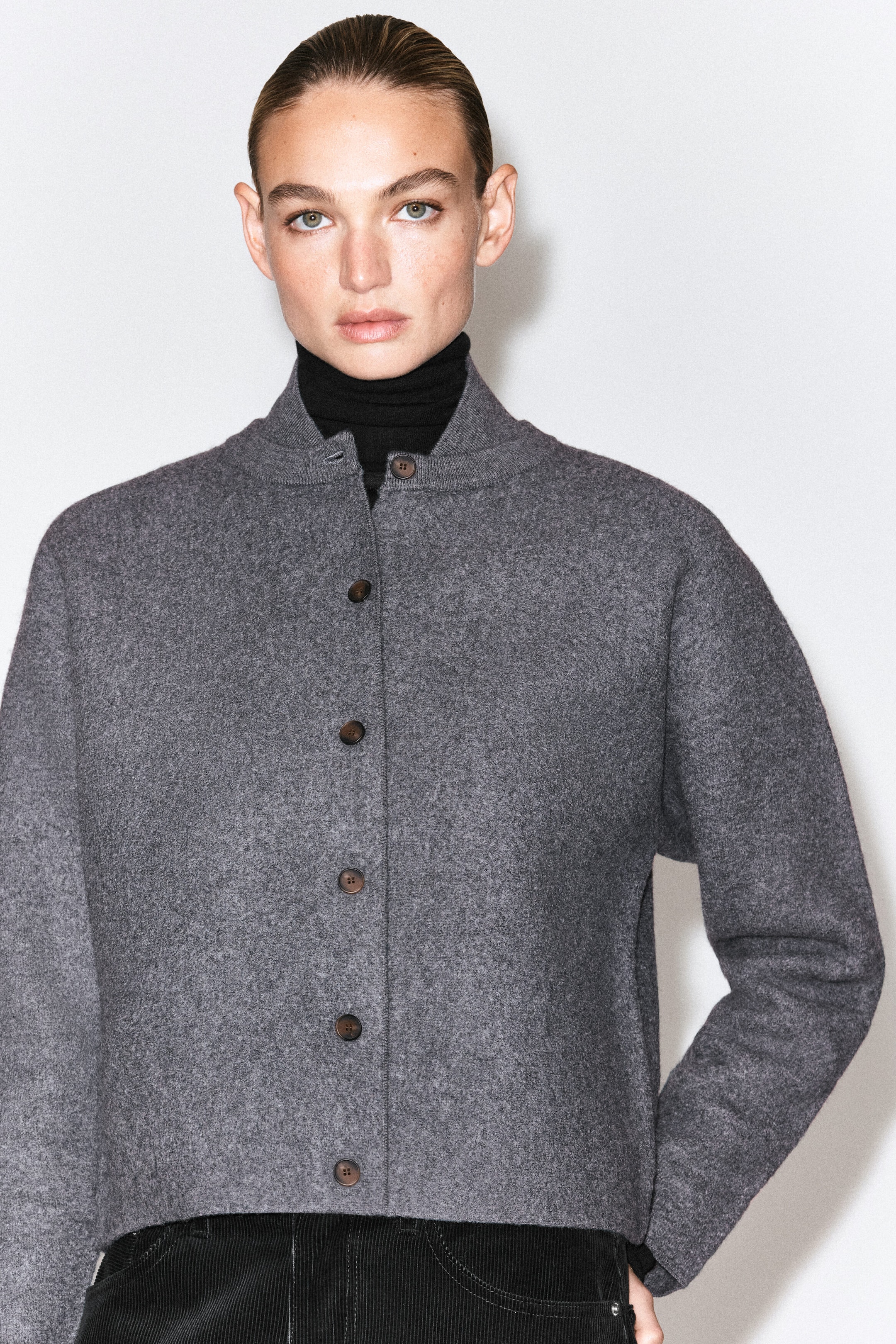 The model is wearing a grey boiled wool cardigan with a ribbed bomber-style collar and front button closure. It is layered over a black turtleneck and styled with black corduroy trousers. The model is wearing a grey boiled wool cardigan with a ribbed bomber-style collar and front button closure. It is layered over a black turtleneck and styled with black corduroy trousers.