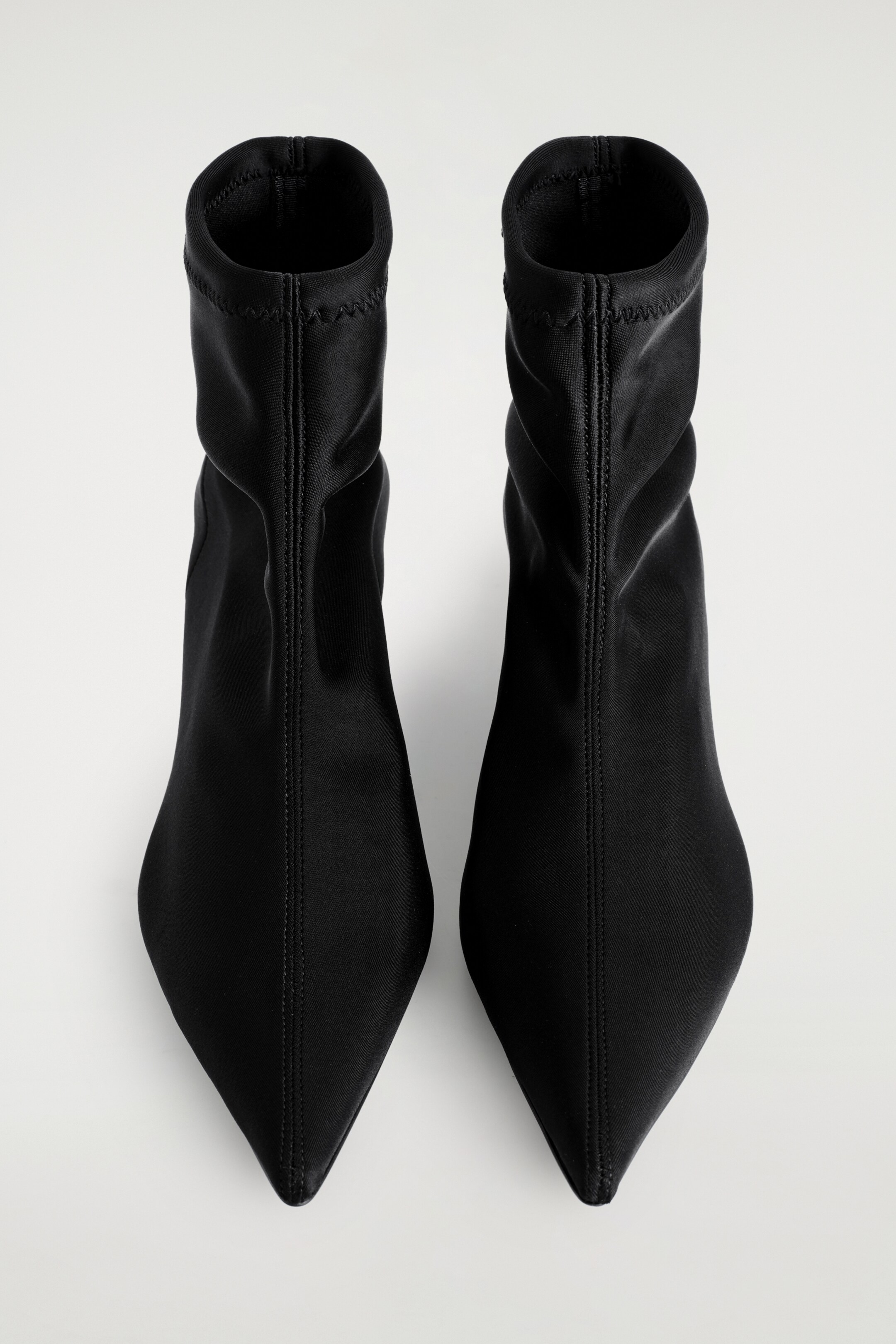 Größeres Bild ansehen: Ein Paar schwarze Sock Boots wird von oben gezeigt, mit schlanken, knöchellangen Schäften und scharf zugespitzten Zehen. Mittige Nähte verlaufen an der Vorderseite jedes Stiefels herunter, und das glatte, matte Obermaterial zeigt subtile Zickzacknähte entlang der elastischen Knöchelmanschetten.