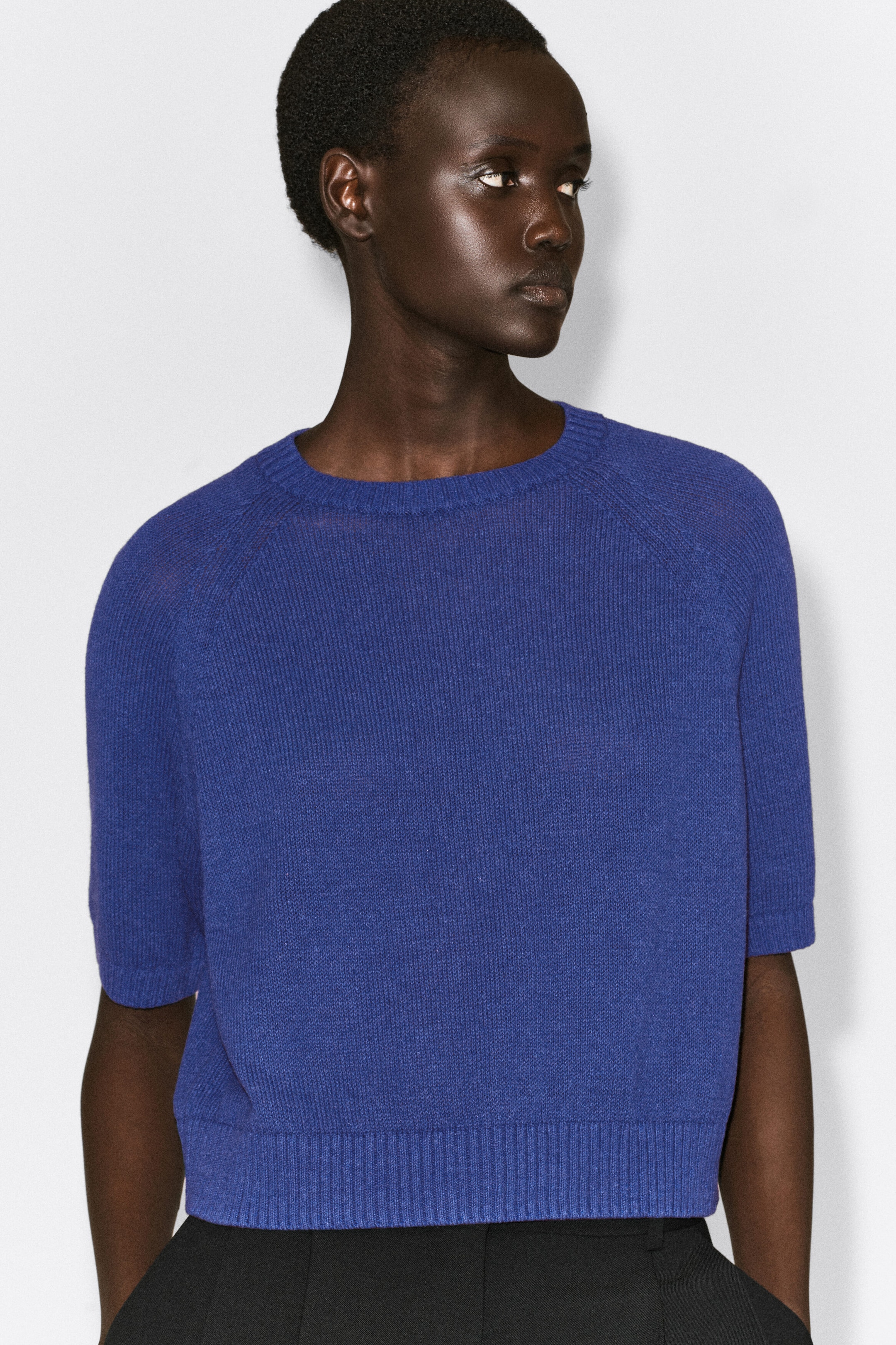 The shrunken knitted linen T-shirt in vibrant blue is shown from the front, highlighting its cropped fit, ribbed crew neck, raglan short sleeves with ribbed cuffs, and wide ribbed hem. The lightweight, finely textured top rests near the waistband of black trousers.