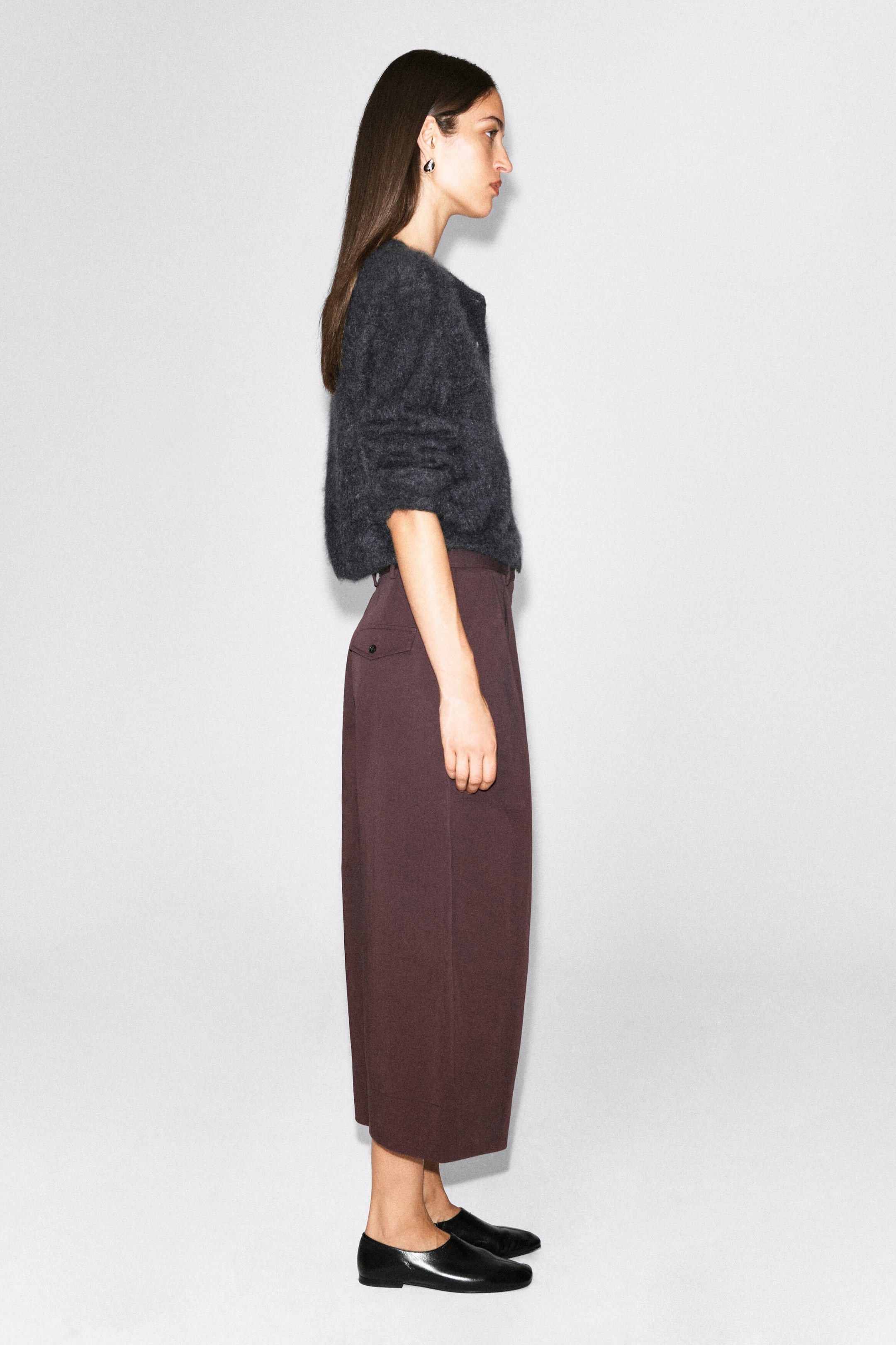 Cotton cropped barrel-leg pants in a dark plum hue are worn by the model, shown in side profile. The pants feature a curved, wide-leg silhouette with a cropped hem and a visible buttoned back pocket, styled with a textured gray knit sweater and black leather flats. Cotton cropped barrel-leg pants in a dark plum hue are worn by the model, shown in side profile. The pants feature a curved, wide-leg silhouette with a cropped hem and a visible buttoned back pocket, styled with a textured gray knit sweater and black leather flats.