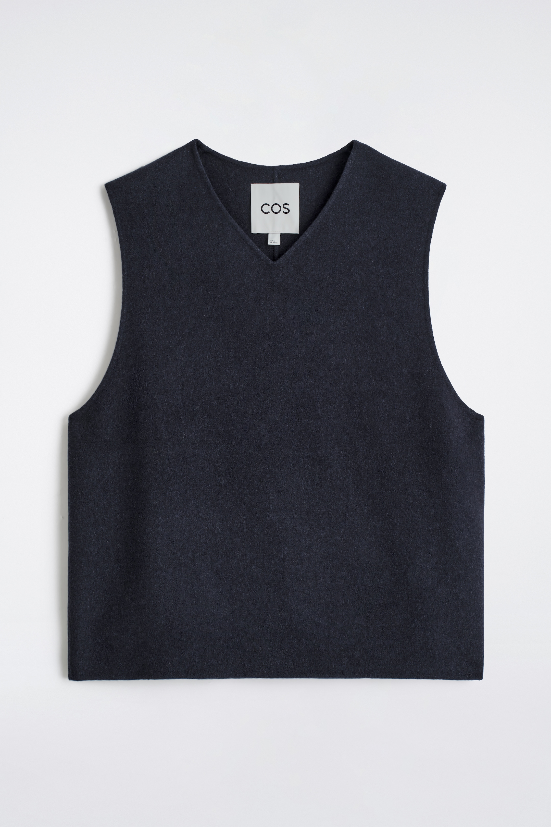 Ver imagen más grande: Un chaleco azul marino de cuello en V se presenta desde el frente, con una silueta relajada y sin mangas que tiene una textura suave y mate. Una etiqueta blanca de la marca COS es visible en el interior del cuello en V.