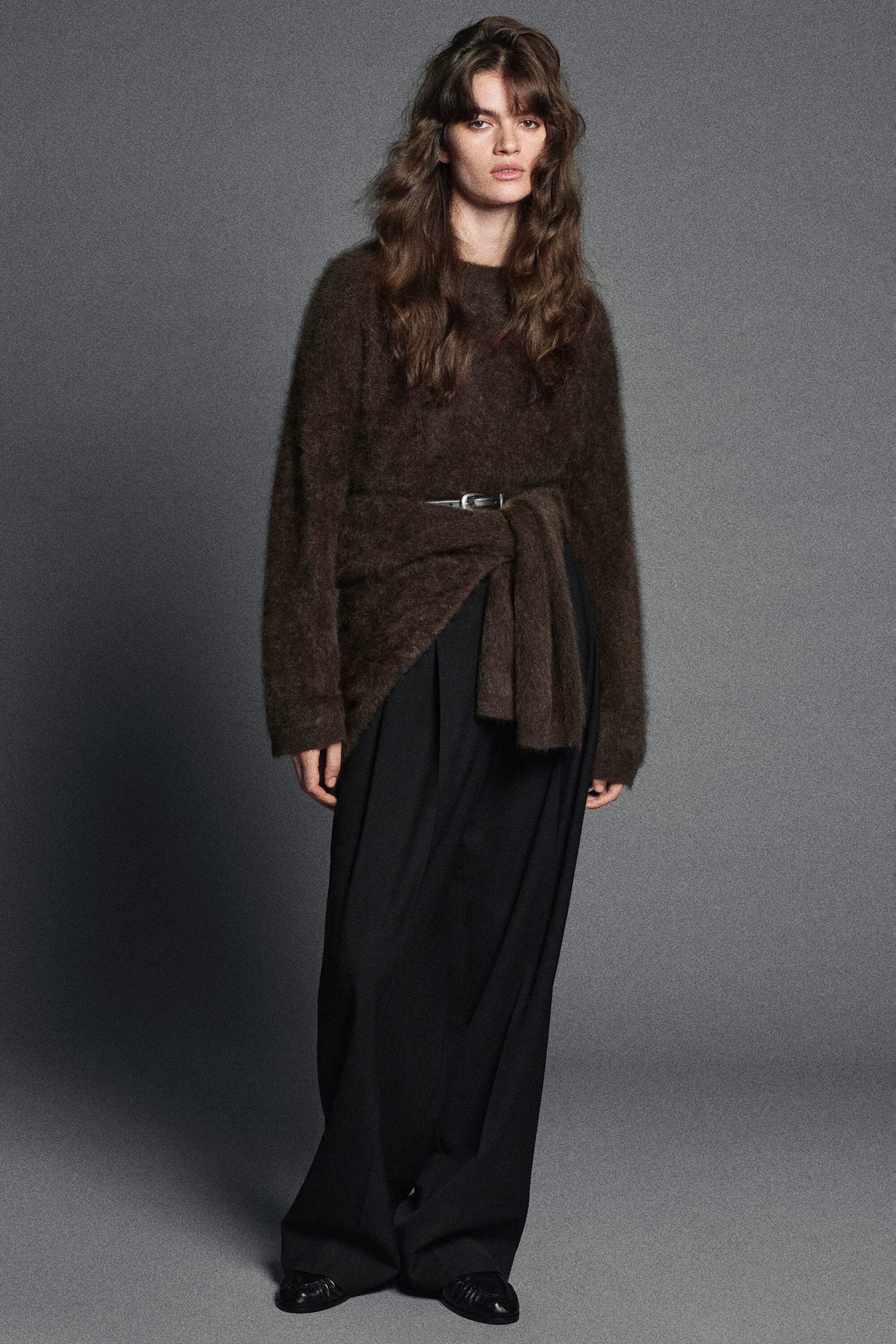 A dark brown, textured crew-neck jumper with a fuzzy, brushed finish is shown from the front. The jumper is styled with a thin black belt cinching the waist to create a draped front and is paired with wide-leg black trousers.