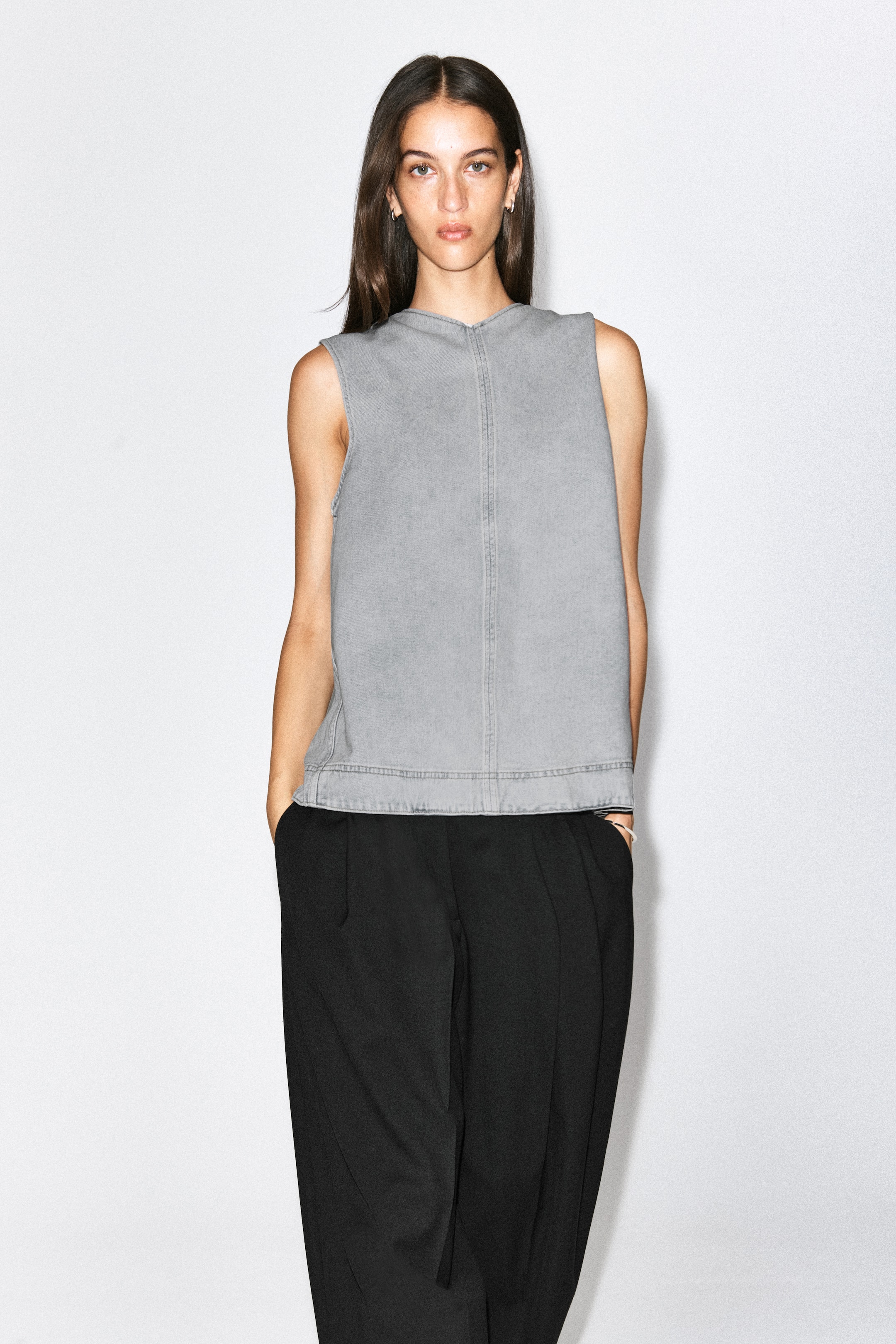 Visualizza l'immagine ingrandita: Una blusa in denim grigio senza maniche, con collo alto e una vistosa cucitura verticale centrale, indossata dalla modella. Il top è abbinato a pantaloni neri dalla vestibilità ampia.
