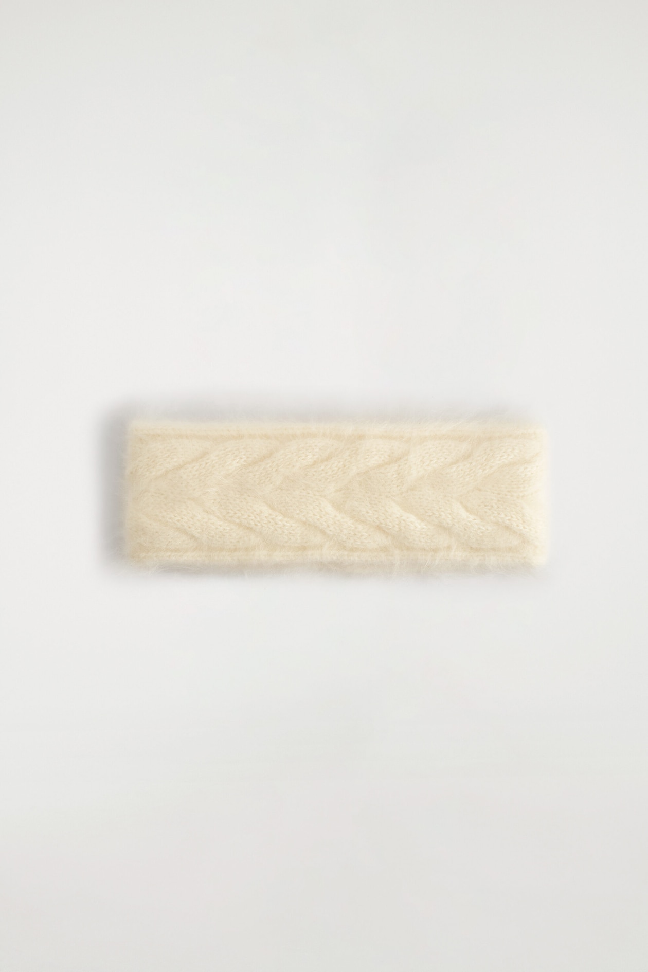 A cream-colored knit headband is lying flat, facing the front to show its central cable-knit pattern. The product features a fuzzy, brushed texture with soft, visible fibers along its edges.