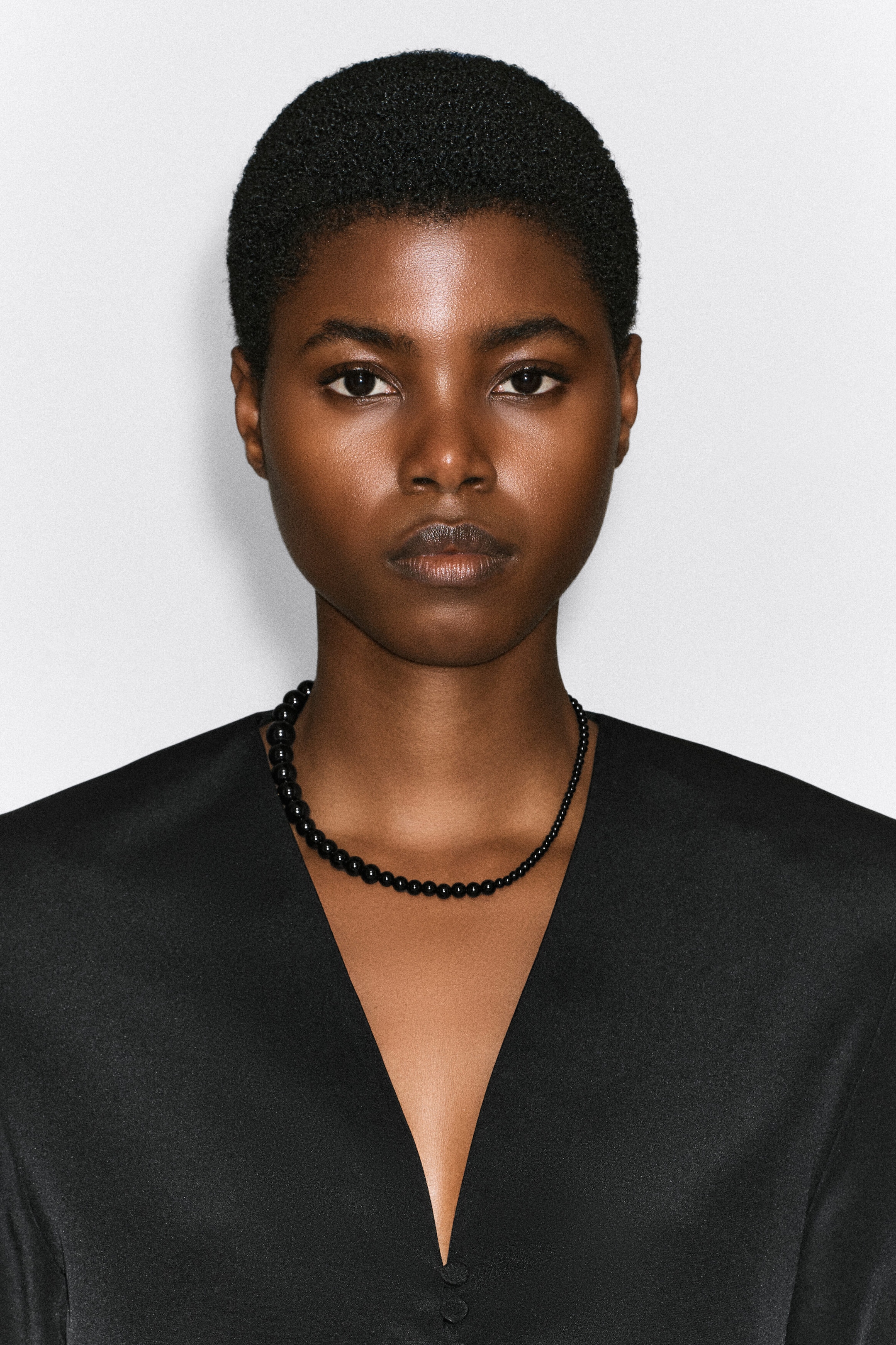 Un collier de perles noires, composé de perles brillantes de taille graduée, est porté autour du cou du mannequin. Le collier est stylisé avec un haut noir à col en V.