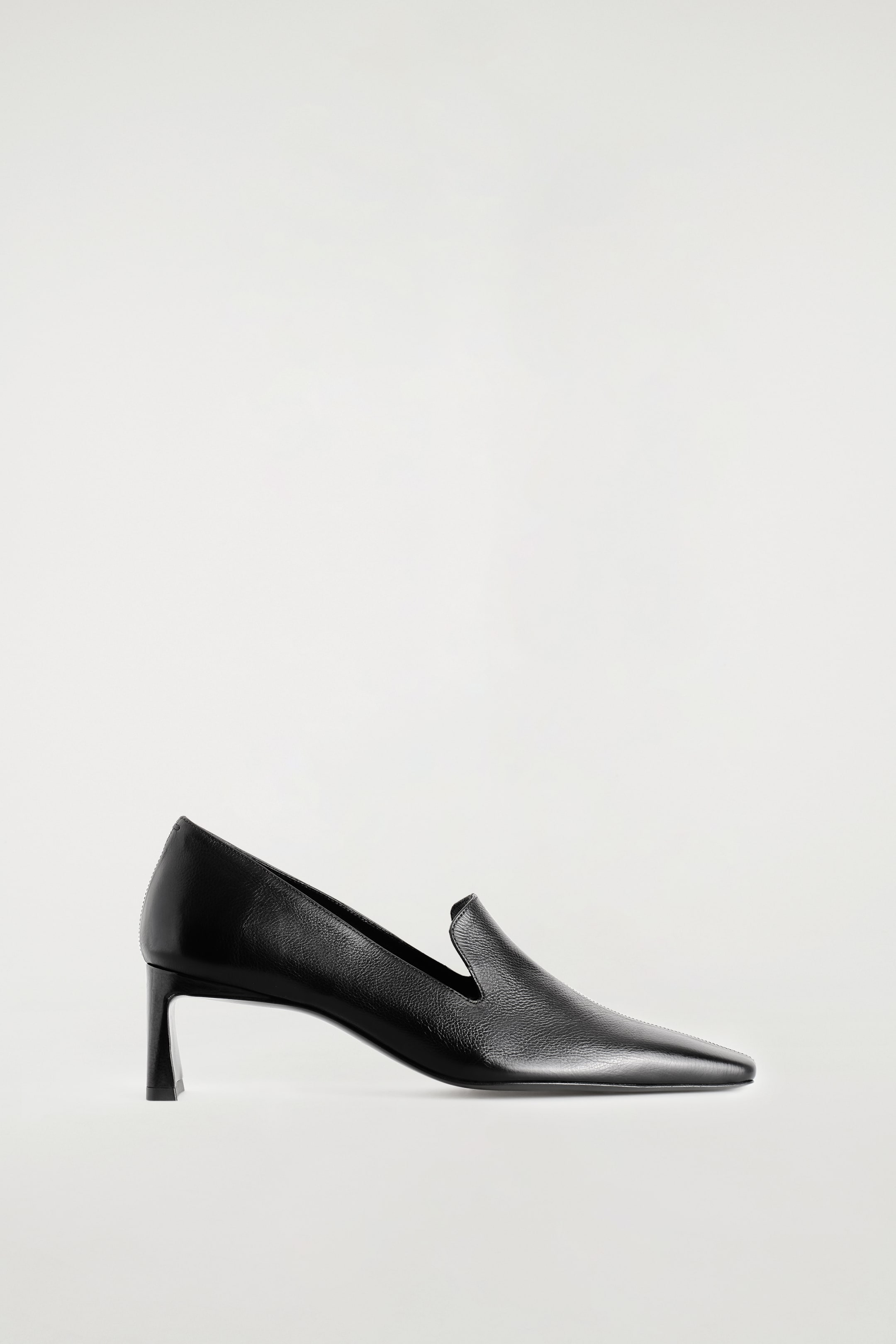 Agrandir l'image: Une chaussure à talon de type mocassin en cuir angulaire présente une finition en cuir noir élégante, vue de profil et orientée vers la droite. Le modèle se distingue par un bout carré net, une empeigne de mocassin à échancrure haute et un talon bas, fin et architectural.
