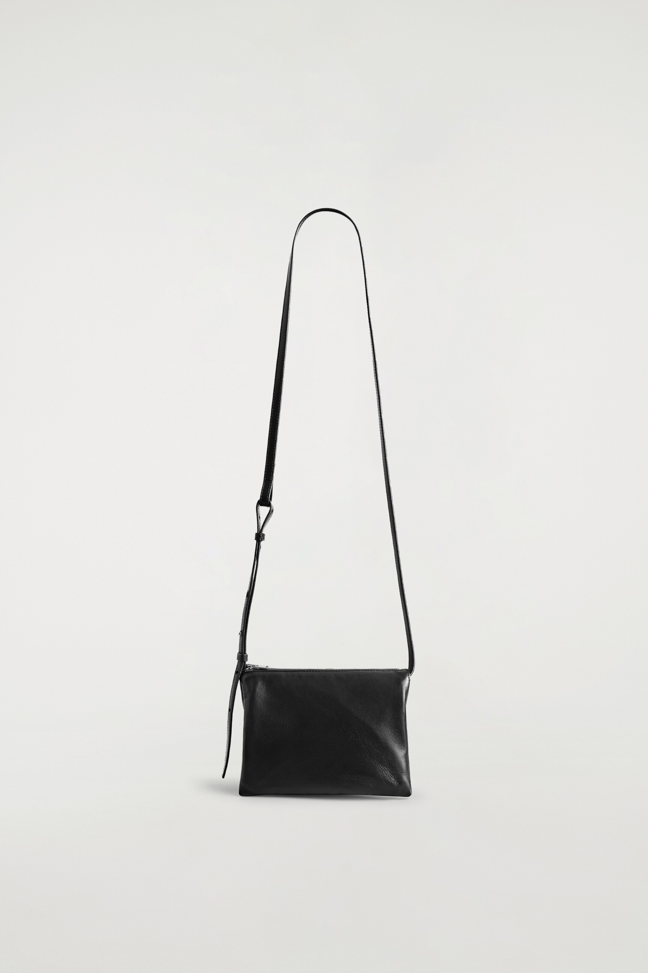 Visualizza immagine più grande: La TROVE CROSSBODY BAG - LEATHER presenta il suo FRONTE, apparendo come una pochette rettangolare realizzata in morbida pelle nera. Una sottile tracolla nera regolabile si estende verso l'alto dagli angoli superiori, e una chiusura con cerniera corre lungo il bordo superiore, con un piccolo tiretto in pelle.