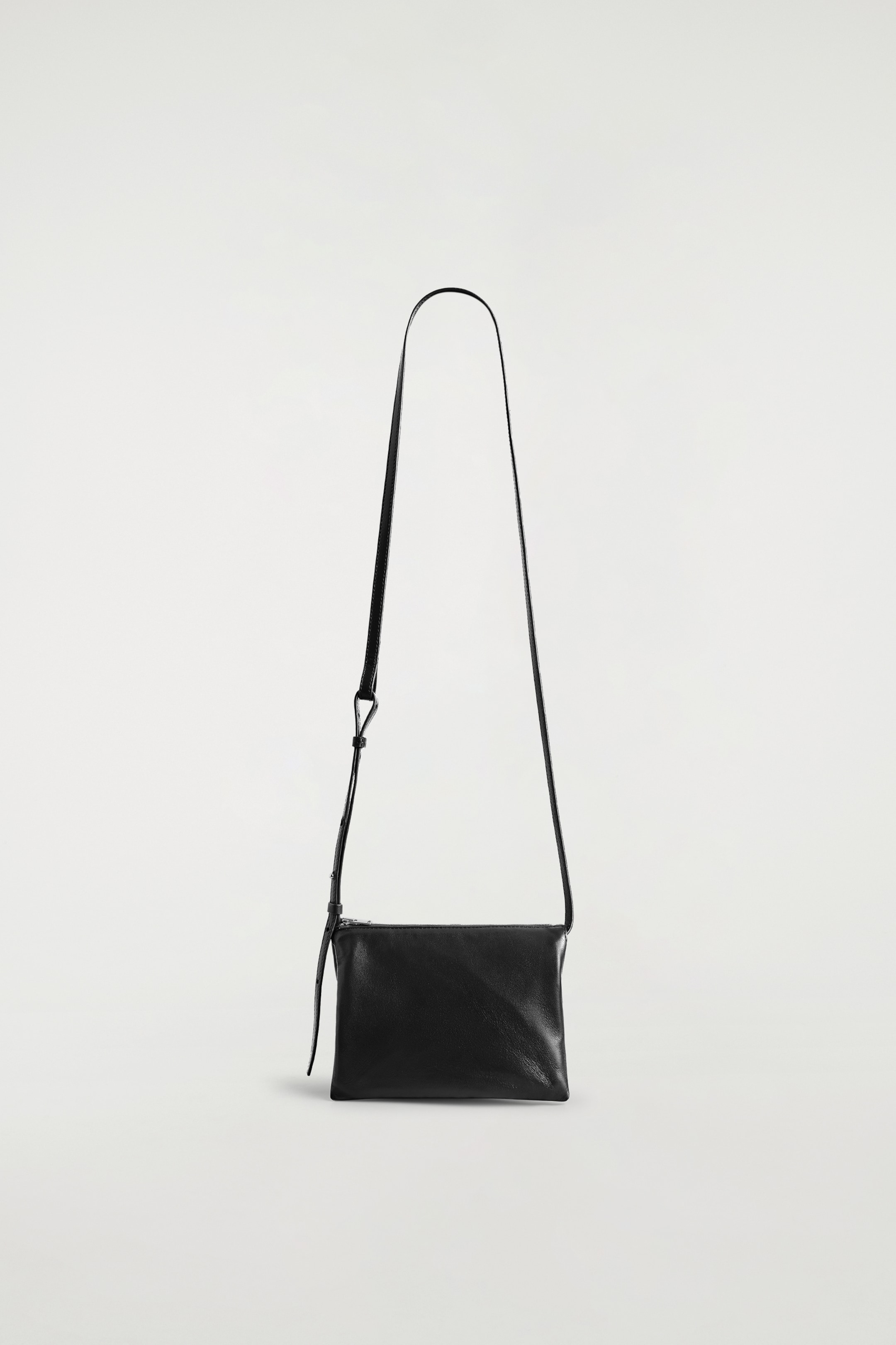 View larger image: The TROVE CROSSBODY BAG - LEATHER is presenting its FRONT, appearing as a rectangular pouch crafted from soft black leather. A thin, adjustable black strap is extending upwards from its top corners, and a zipper closure is running along the top edge, featuring a small leather pull tab.