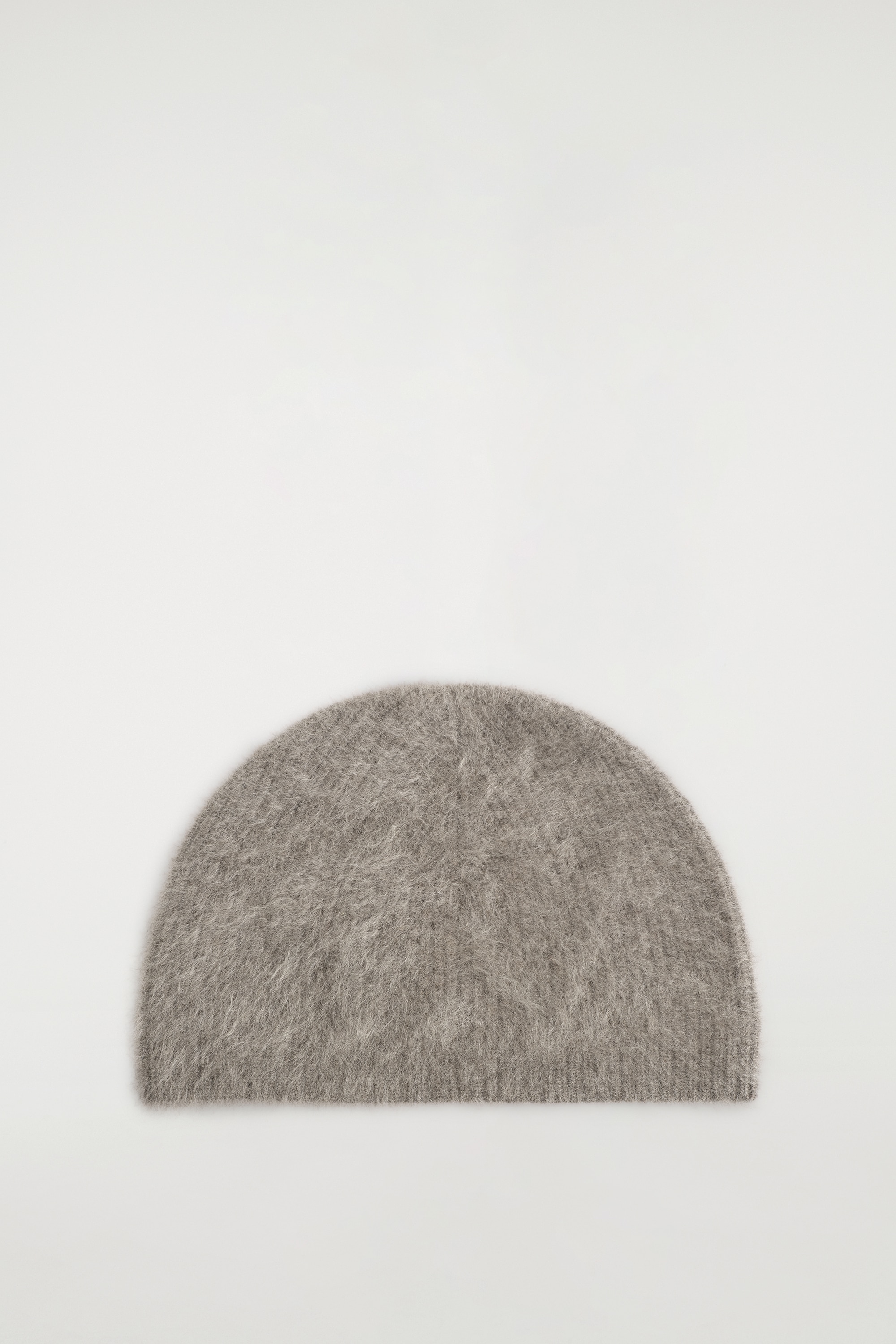 En ljusgrÃ¥ mÃ¶ssa i borstad kashmir bÃ¤rs av modellen och tÃ¤cker huvudet ner till pannan. MÃ¶ssan har en fluffig, texturerad yta med ribbad kant och kombineras med en mÃ¶rkgrÃ¥ texturerad kappa.
