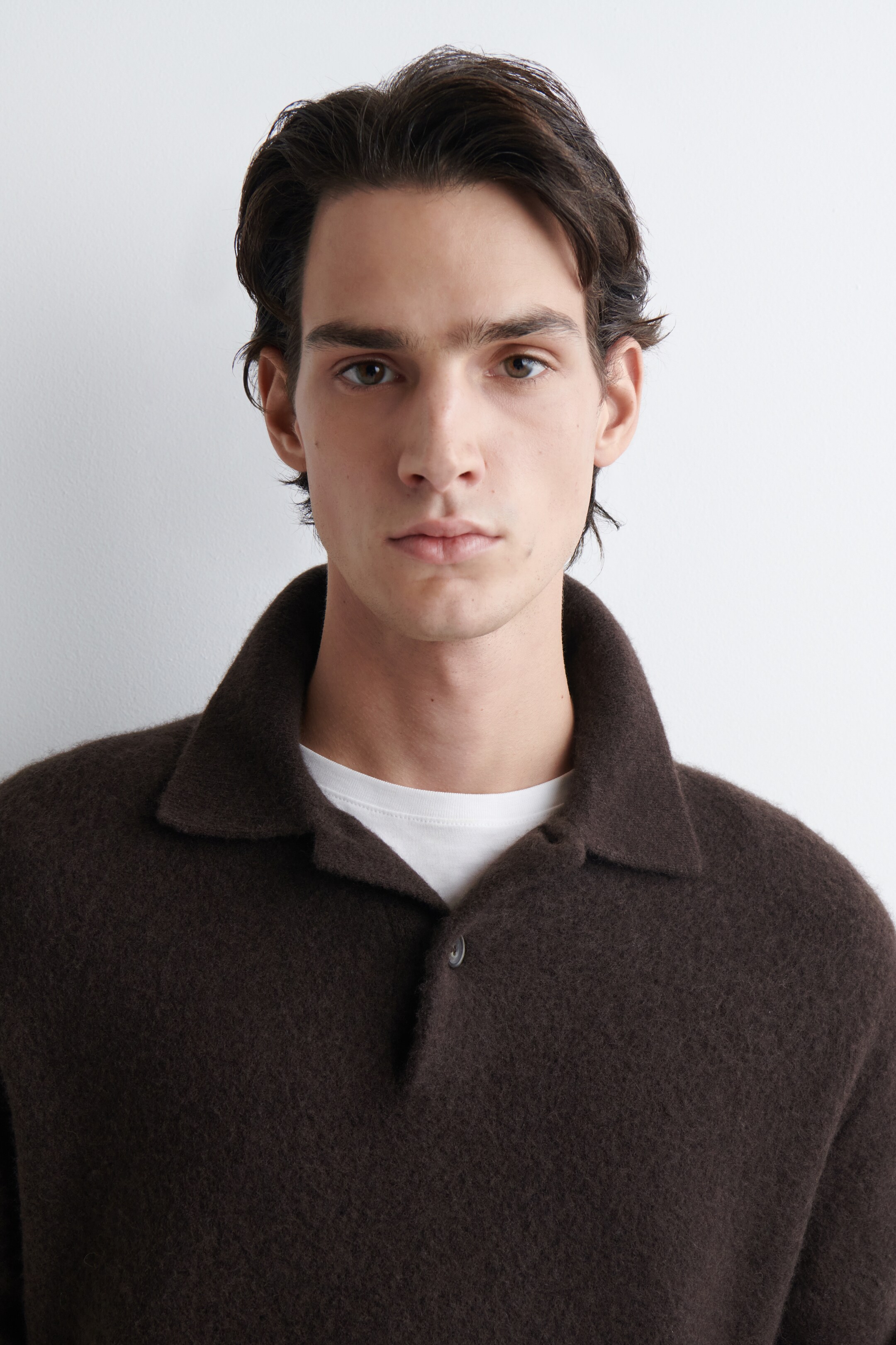 Agrandir l'image: Un pull de style polo marron foncé est montré de face, présentant une texture douce et duveteuse et une patte à deux boutons. Un t-shirt blanc à col ras du cou est visible sous le col.